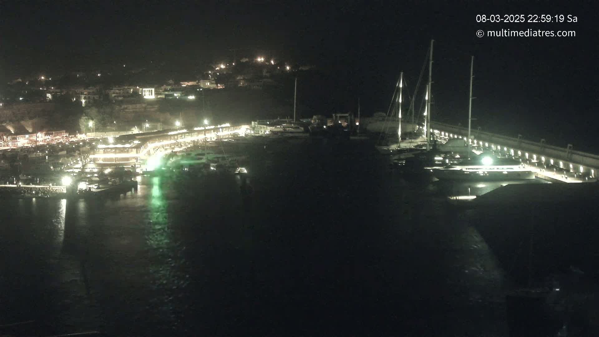 A nighttime view of a harbor filled with boats docked alongside a lit waterfront, with a hillside town visible in the background under a clear, dark sky.