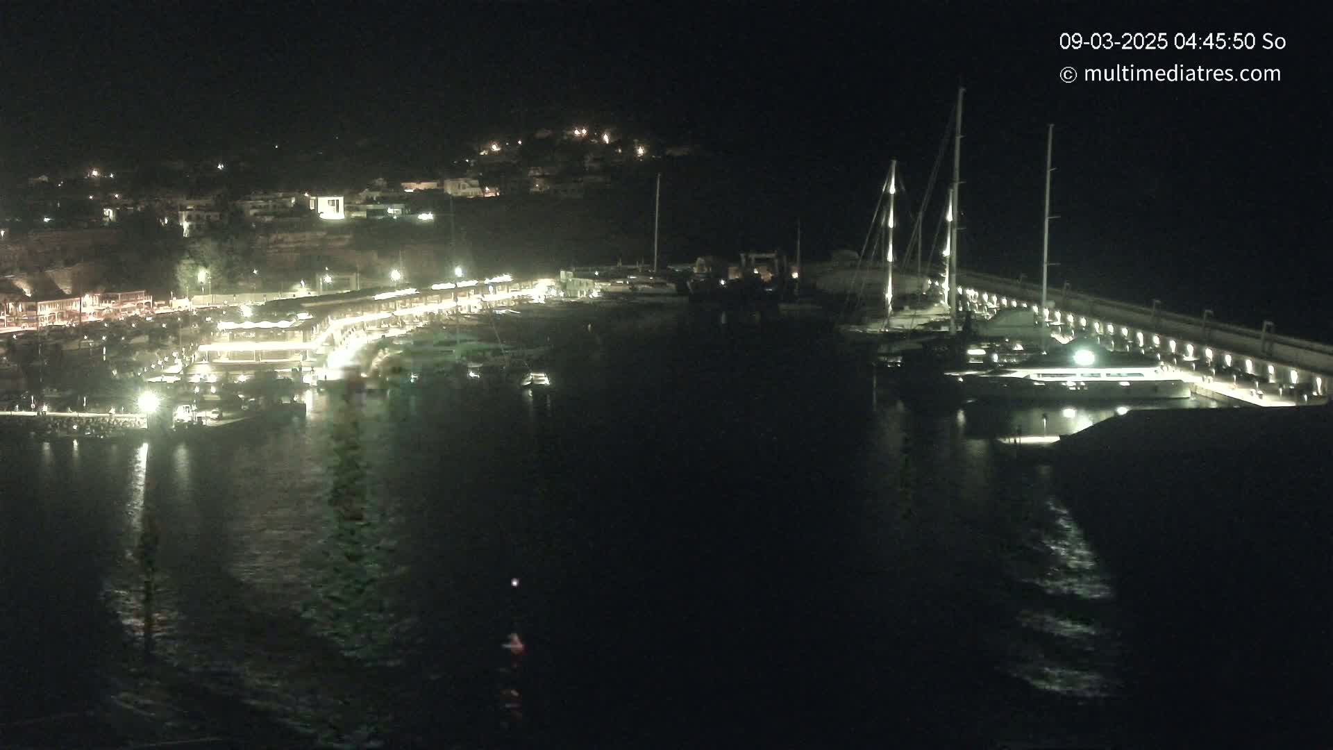 A marina at night, illuminated with lights, showing several boats docked and a town in the background.