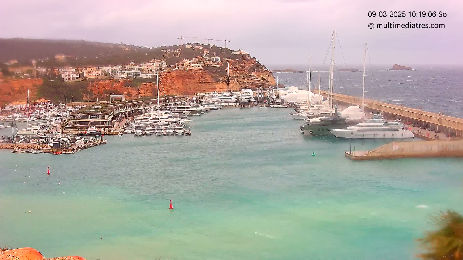 A marina filled with numerous yachts and boats is situated next to a cliffside town under an overcast sky, with choppy water indicating windy conditions.