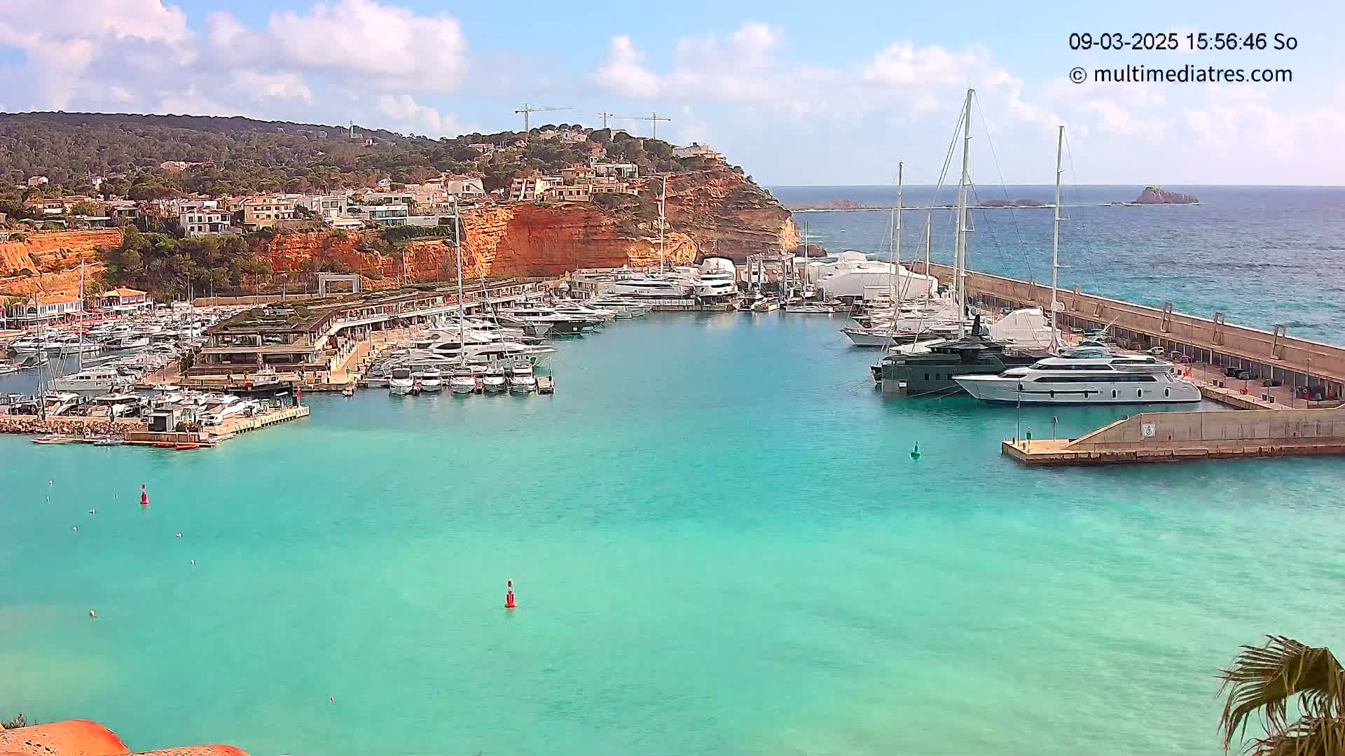 A marina filled with numerous yachts and boats is nestled in a calm, turquoise bay under a partly cloudy sky, with a hillside town and construction cranes visible in the background.