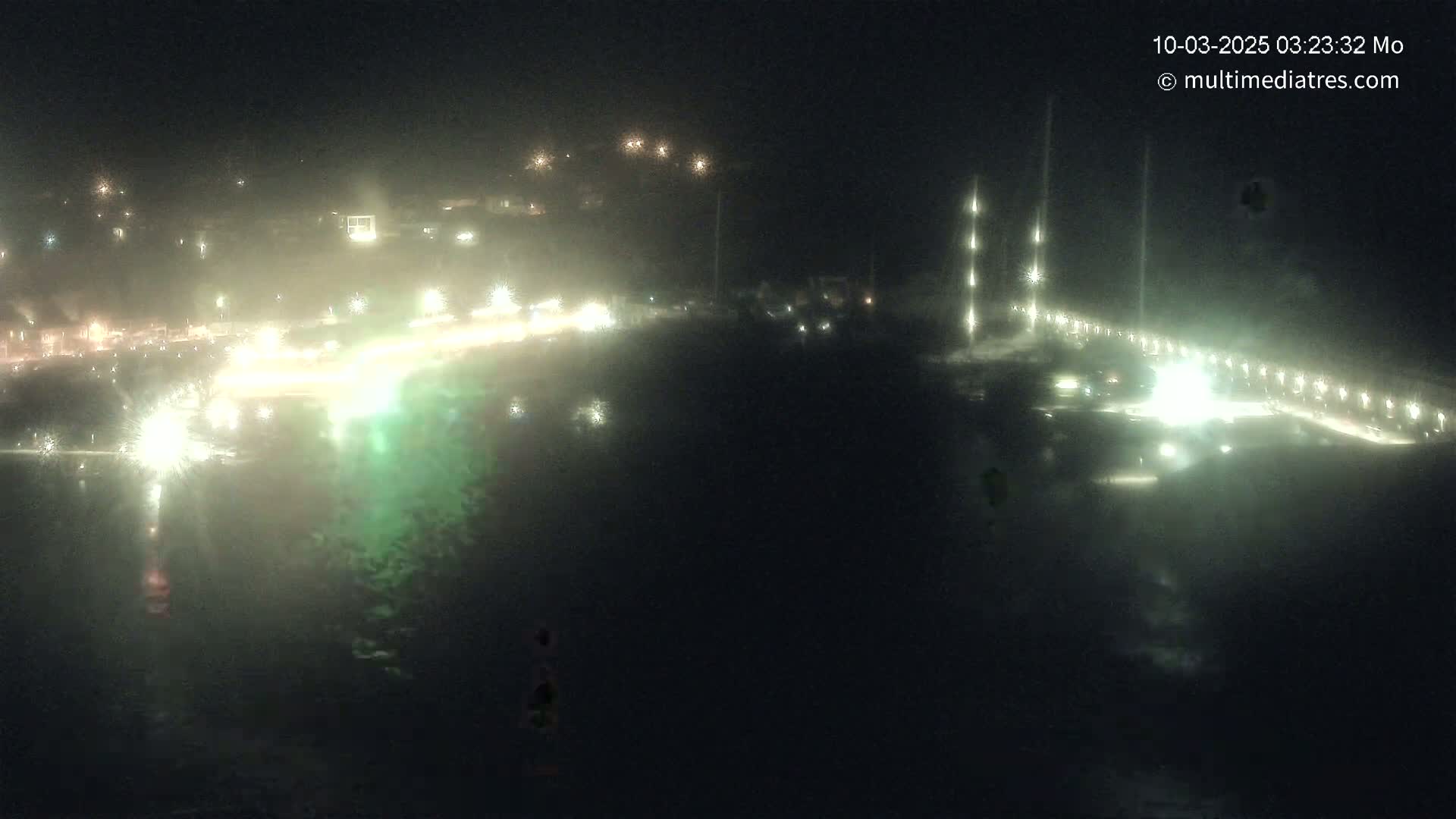 A nighttime, hazy view of a coastline with illuminated buildings and a pier extending into dark water.