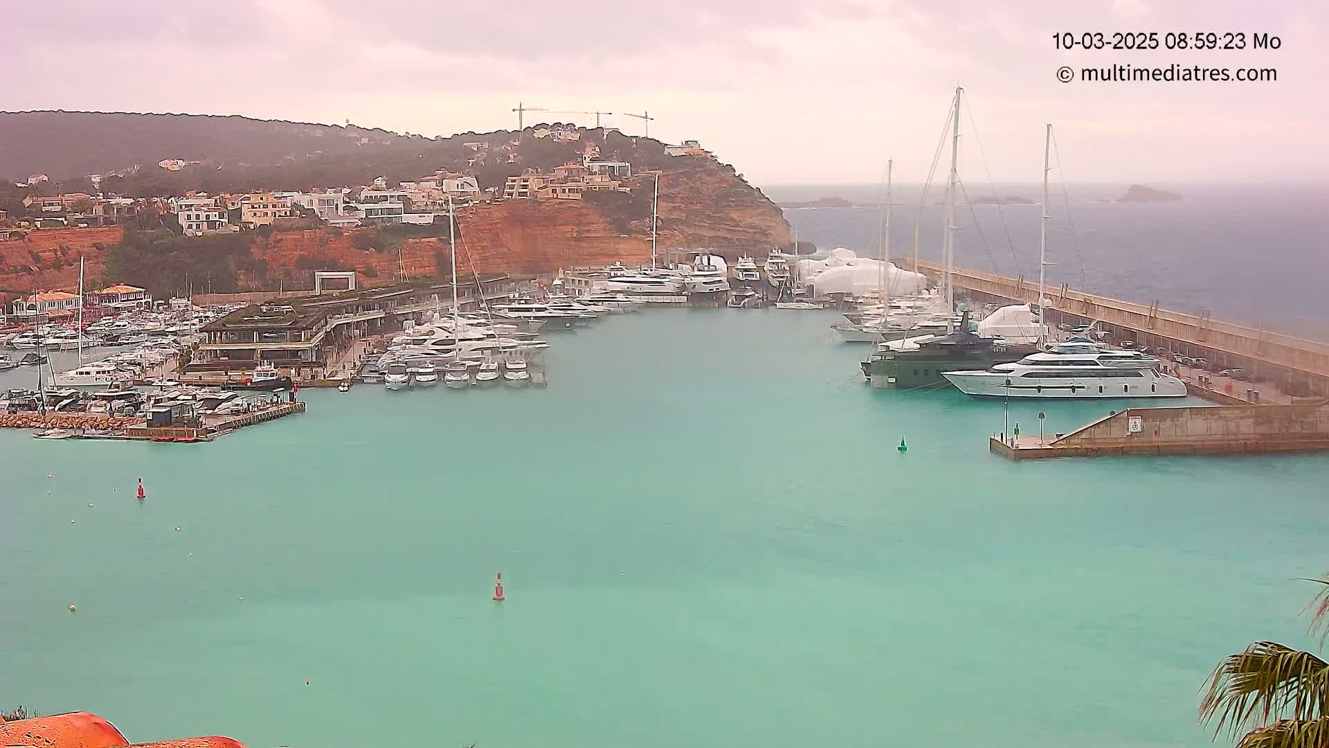 A marina filled with numerous yachts and boats is situated in a calm, teal-colored harbor, under an overcast sky.
