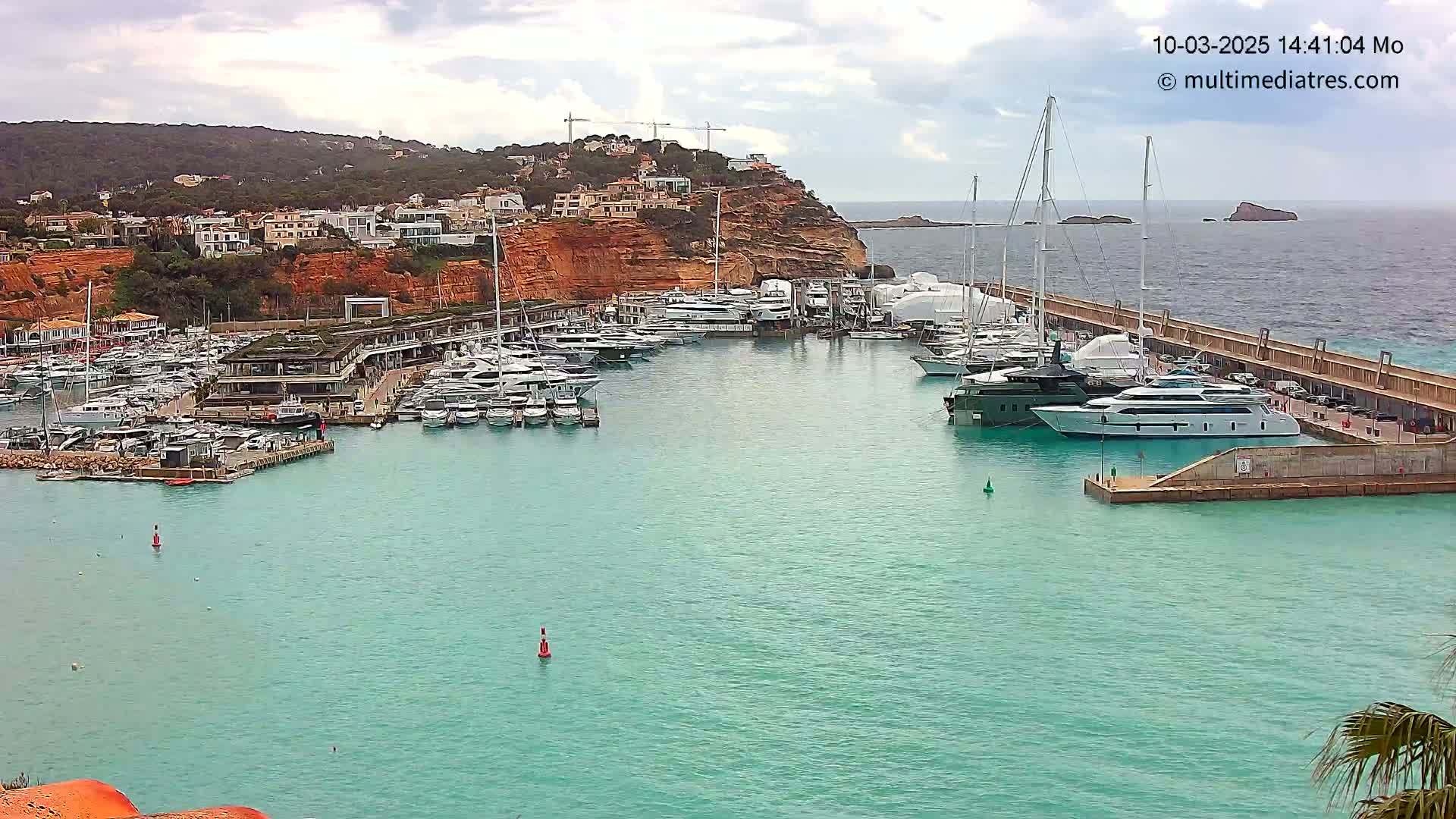 A marina filled with numerous yachts and boats is situated in a calm, turquoise bay, with a hillside town and overcast skies in the background.