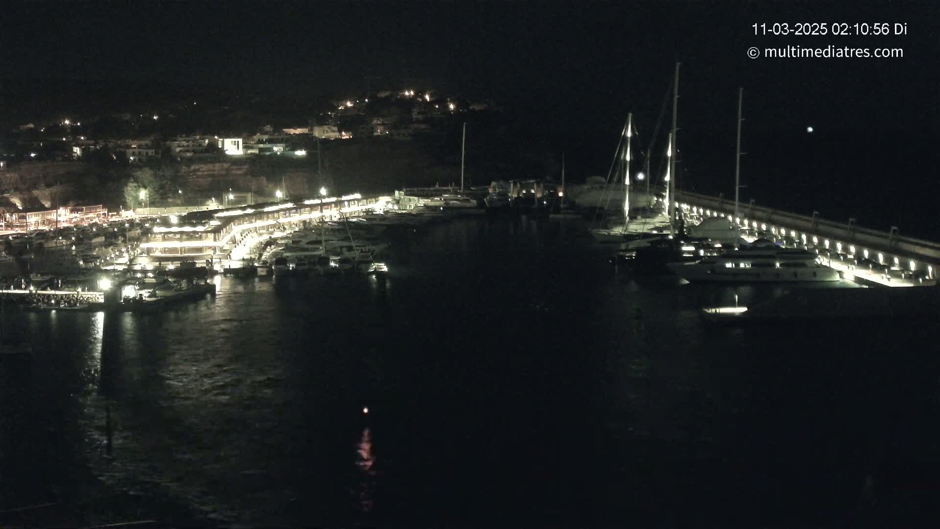 A nighttime view of a marina filled with boats, docked along a lit pier, with a town visible in the background.