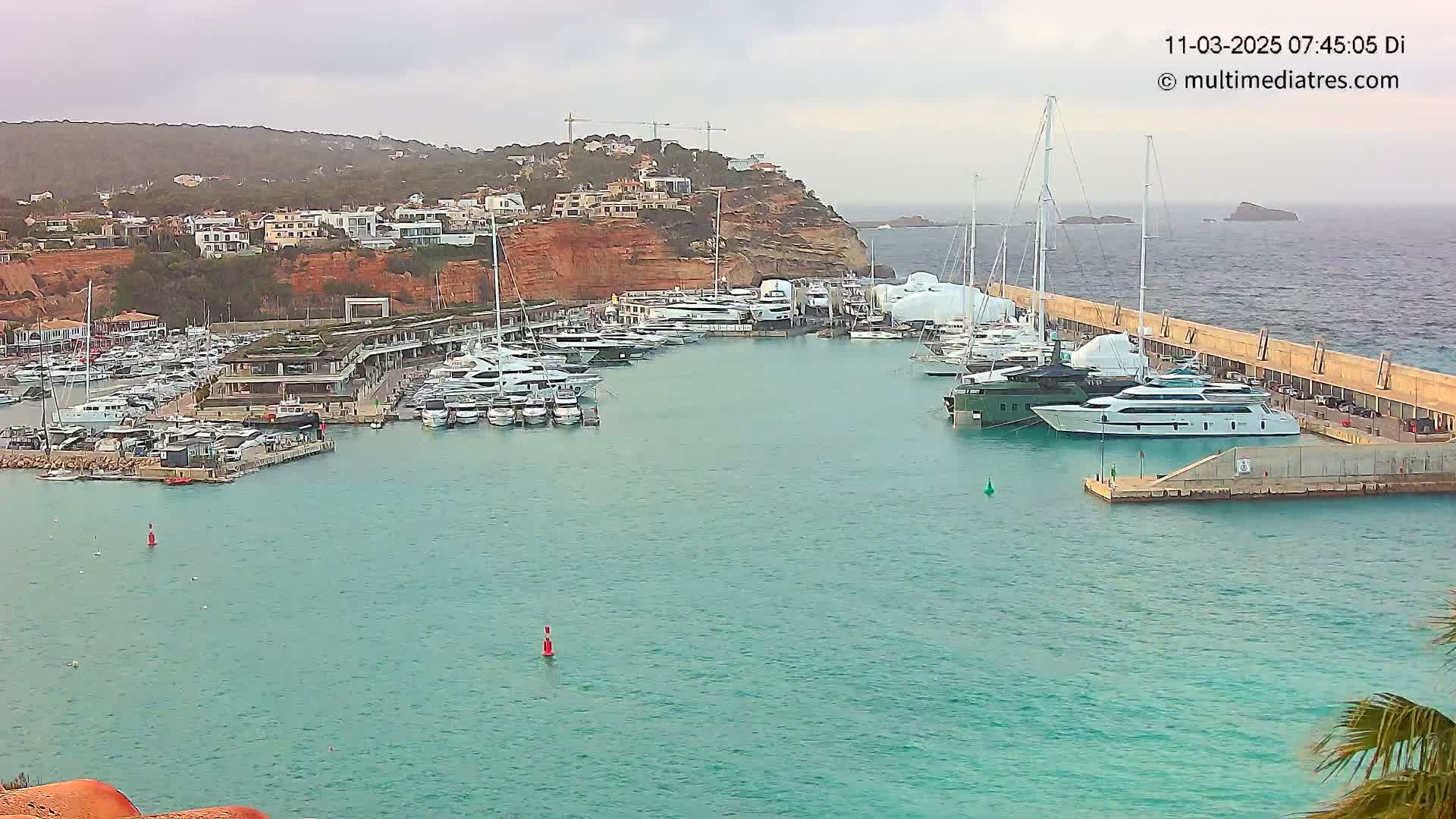 A marina filled with numerous yachts and boats is situated along a calm, turquoise body of water next to a red-cliffed hillside under an overcast sky.