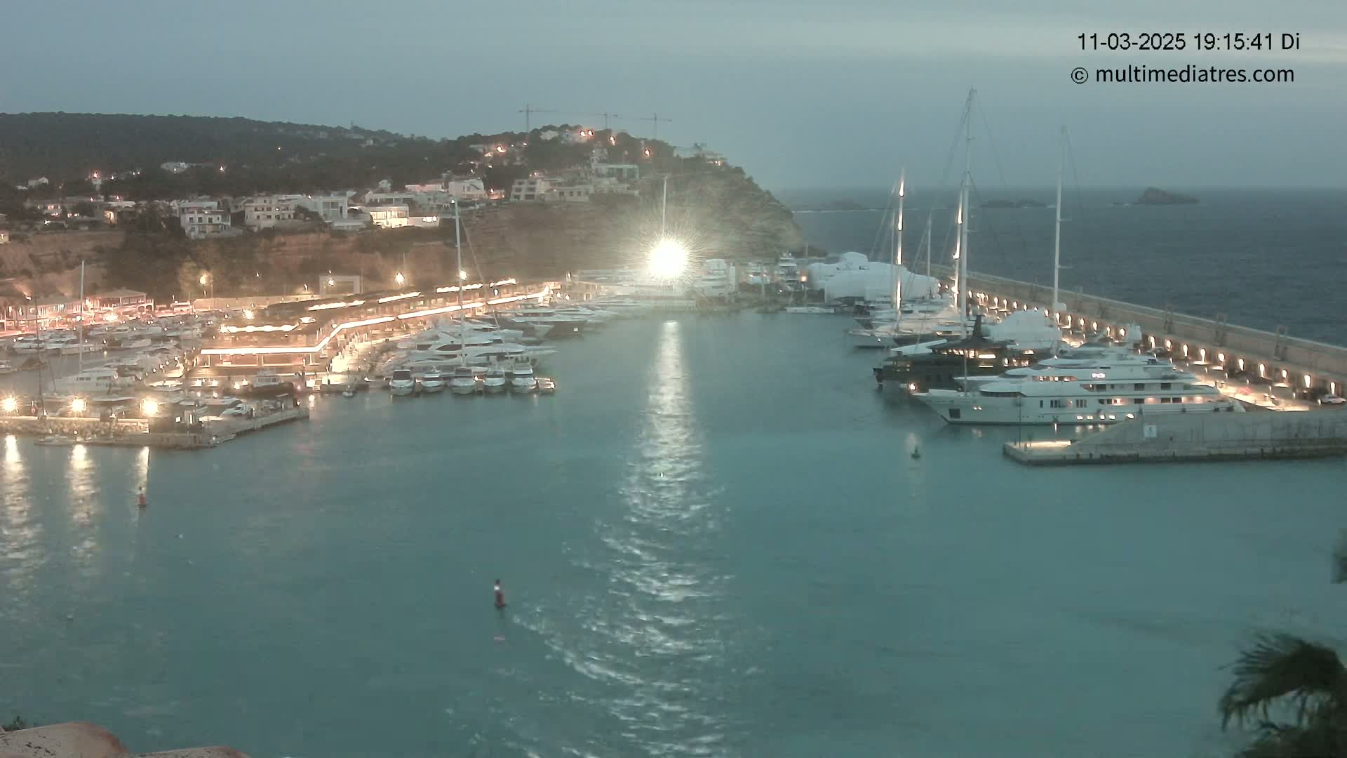 A marina filled with numerous yachts and boats at night, under a dusky sky, is illuminated by lights from the buildings and docks.