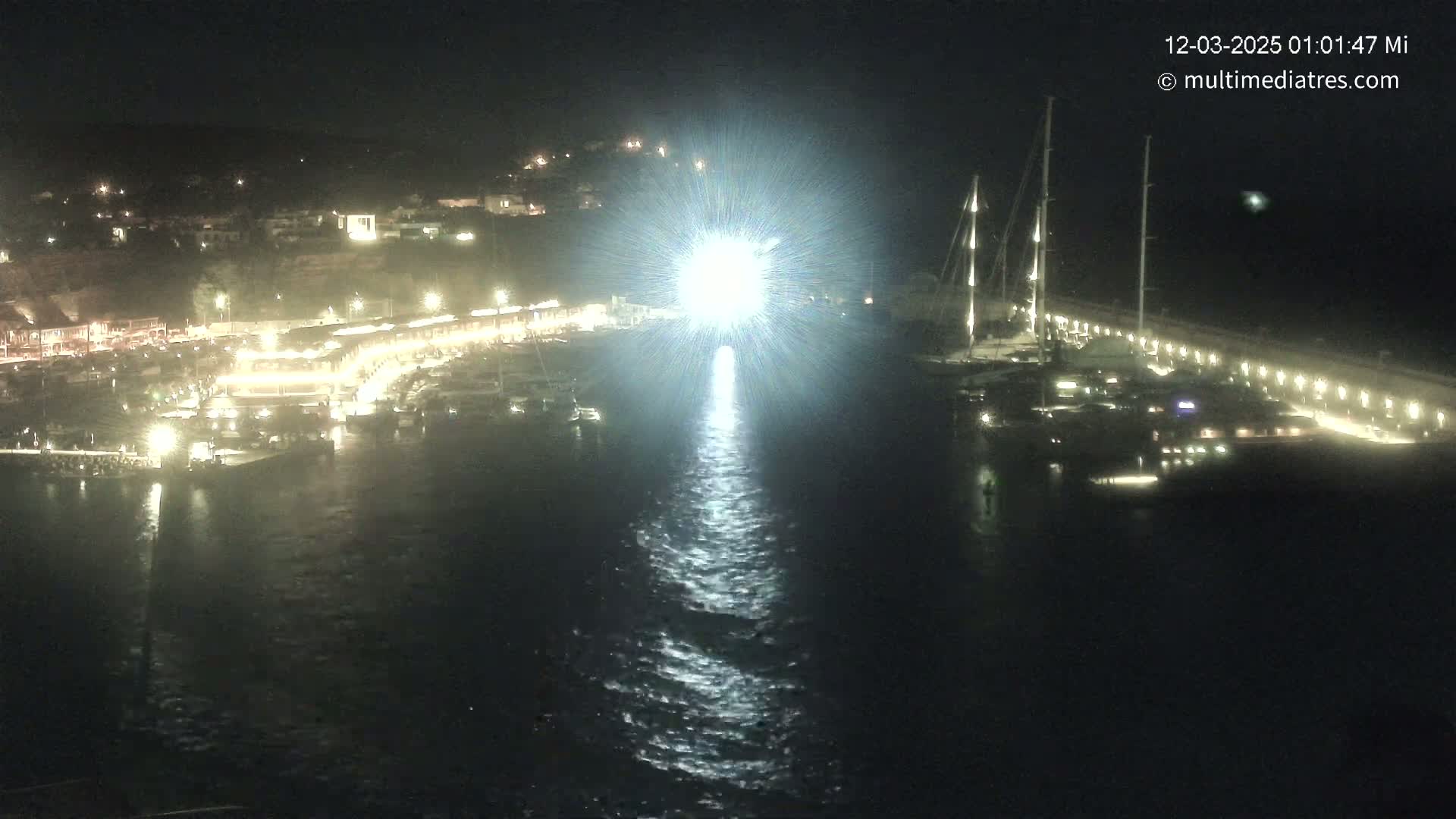 A brightly lit harbor at night, filled with boats and a bright light reflecting on the water.