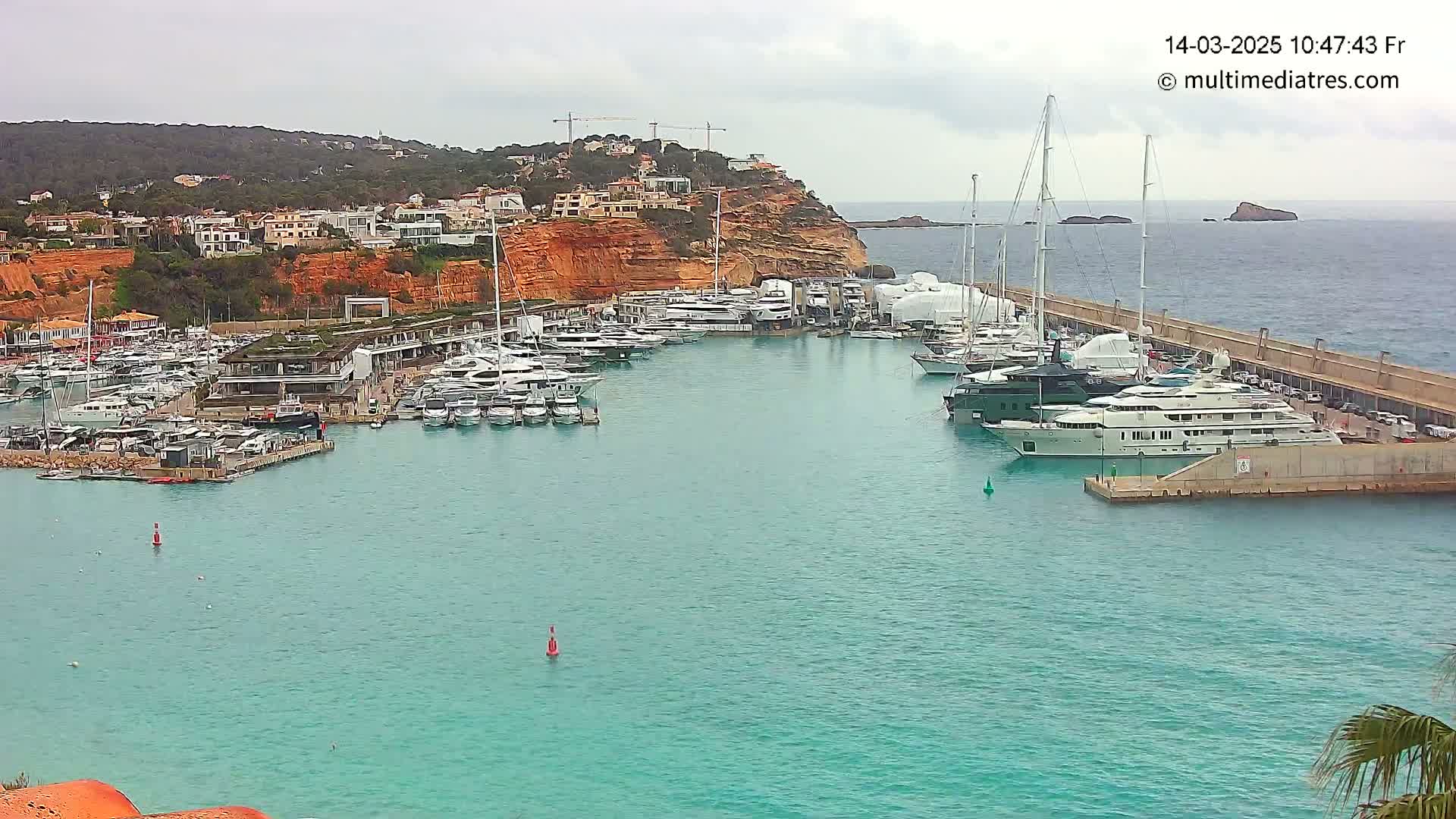 A marina filled with numerous yachts and boats is situated along a coastline under an overcast sky.