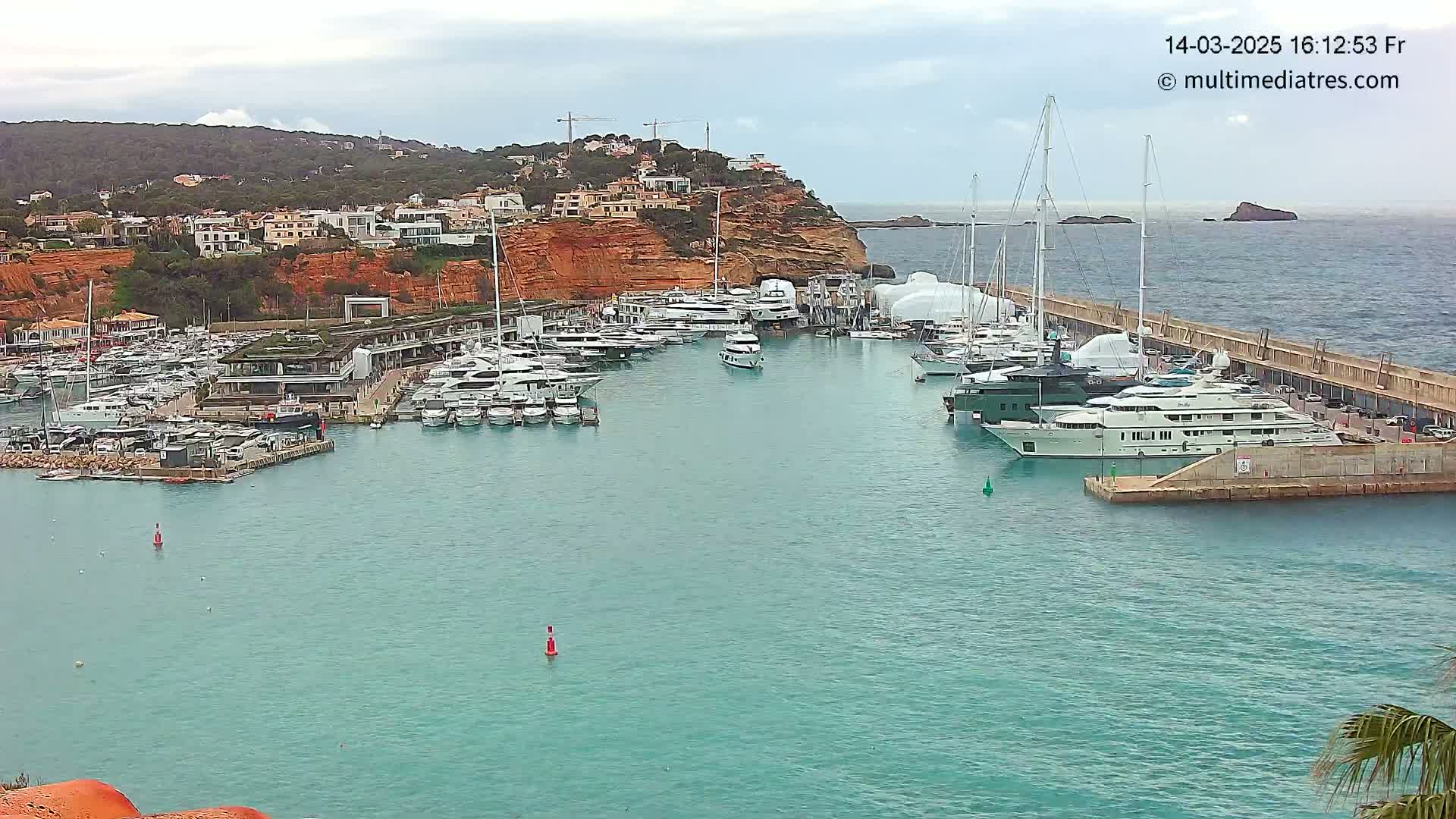 A marina filled with numerous yachts and boats is nestled against a hillside of red-toned buildings under an overcast sky.