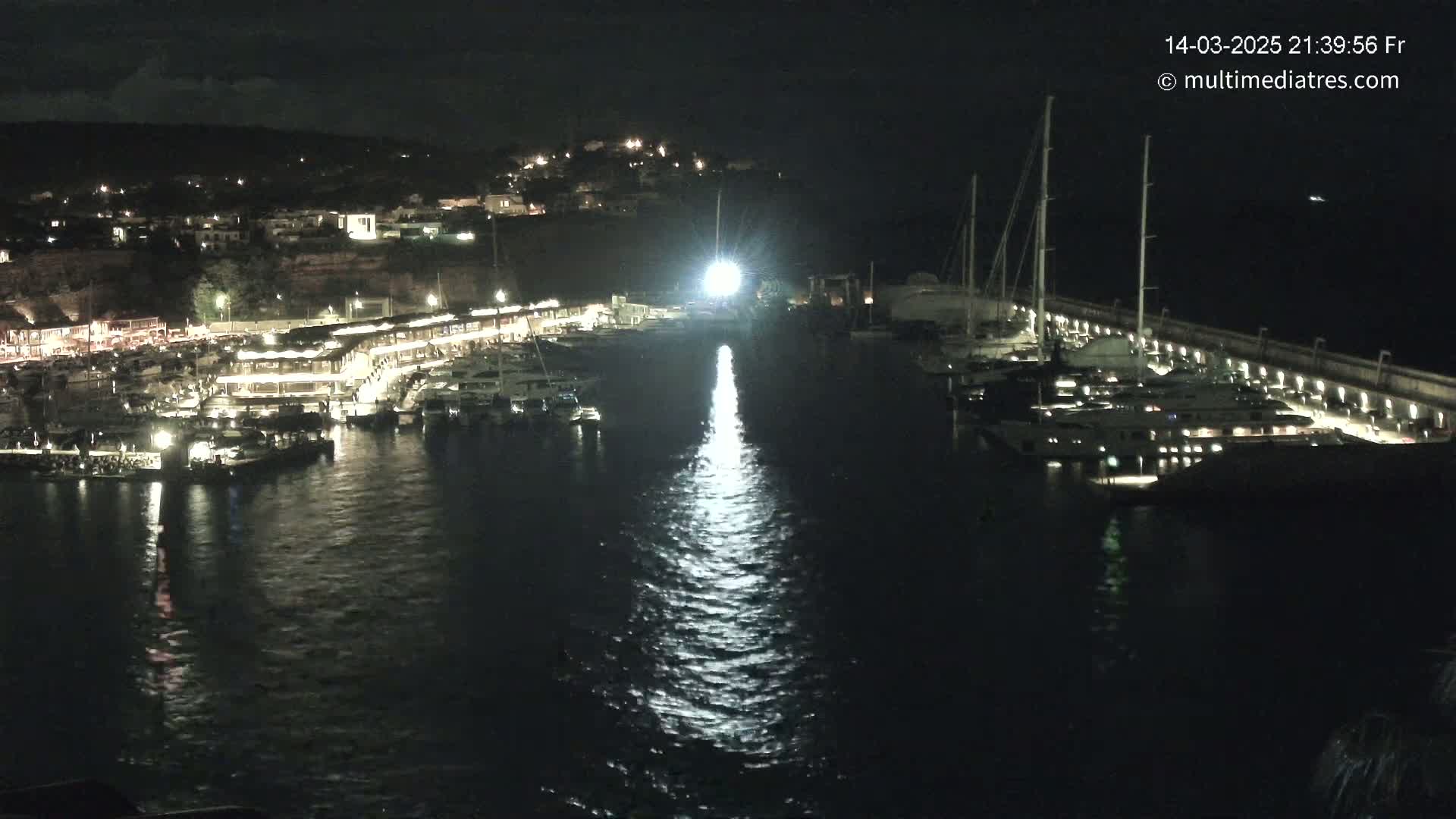 A marina at night, filled with docked boats and illuminated by lights, reflecting on the calm water.