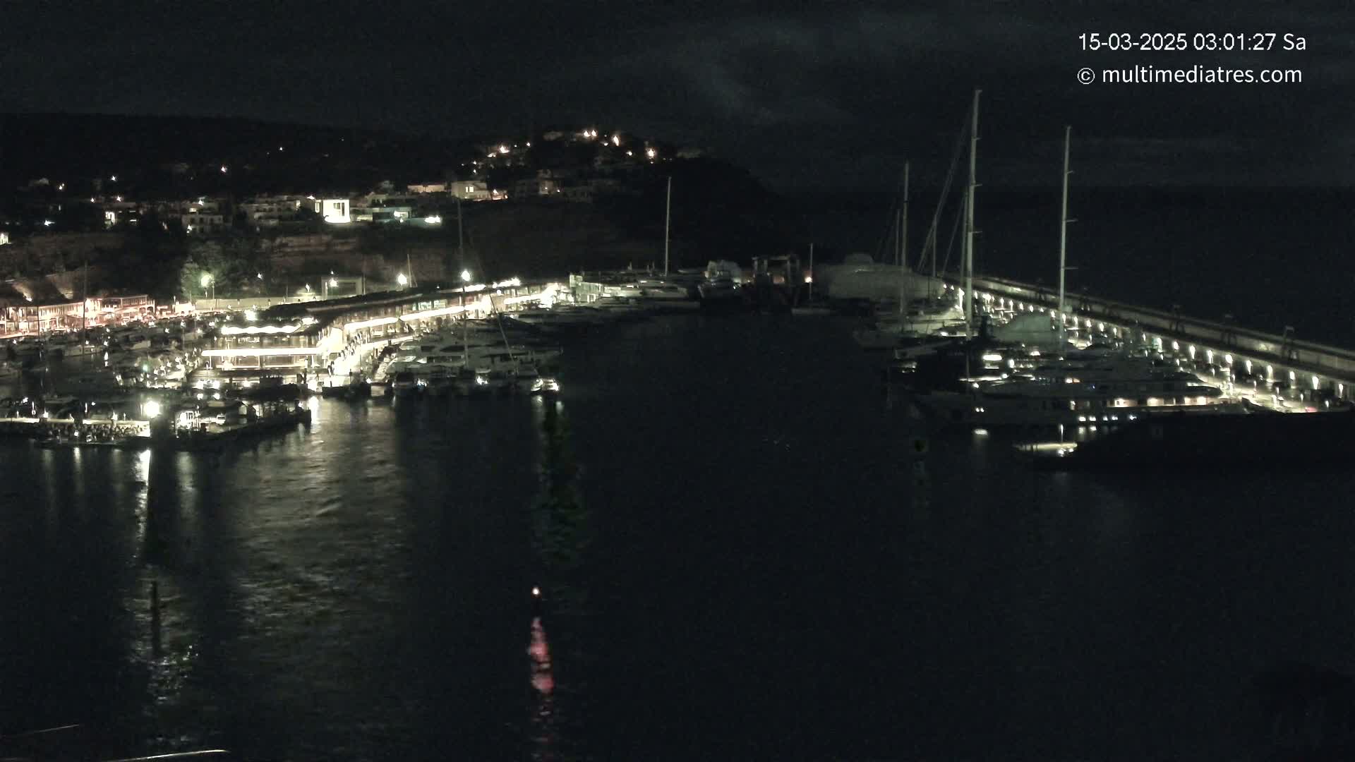 A nighttime view of a marina filled with boats, with a hilltop town lit up in the background under a partly cloudy sky.