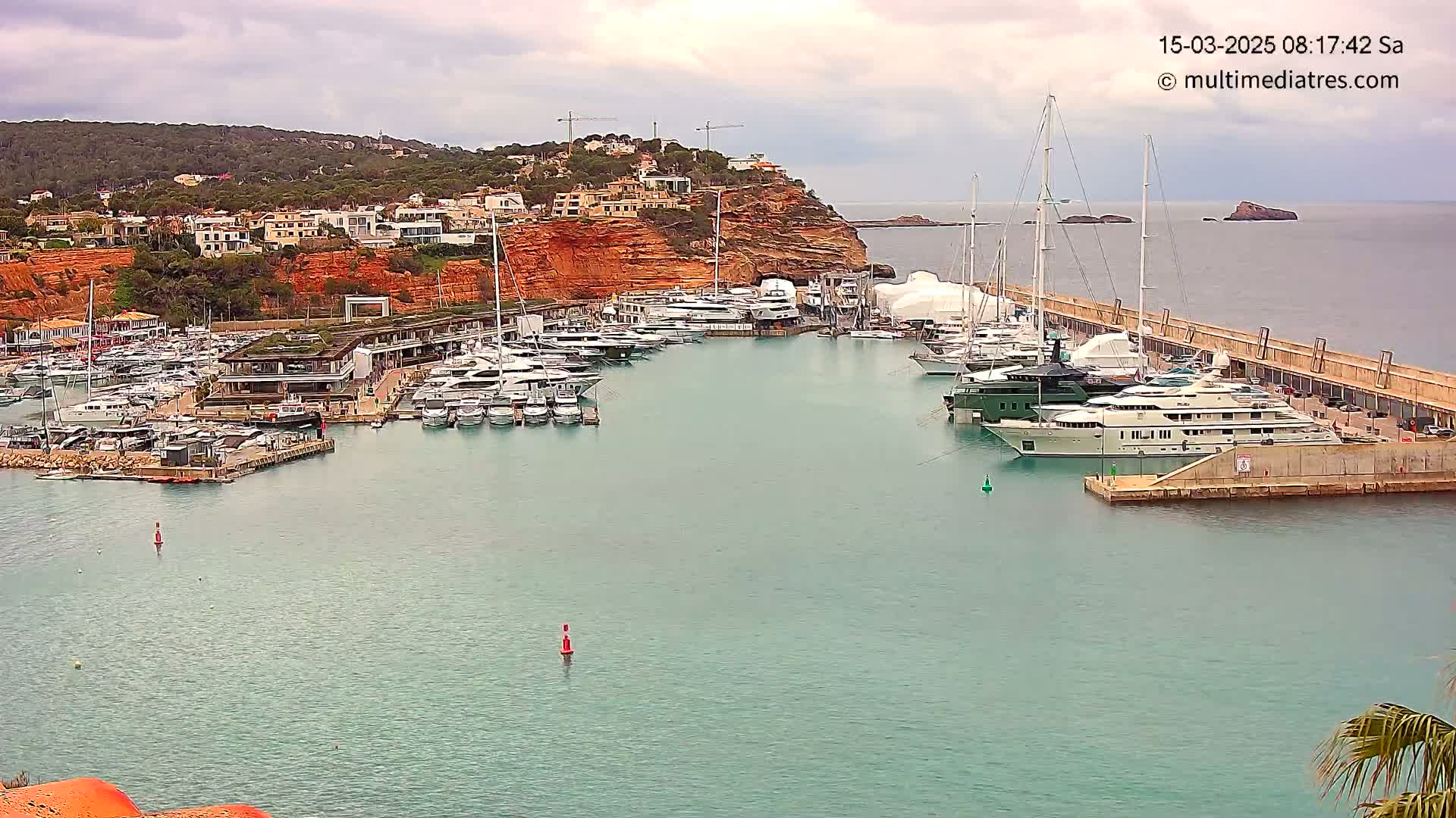 A marina filled with numerous yachts and boats is situated next to a hillside of red-toned buildings under an overcast sky.