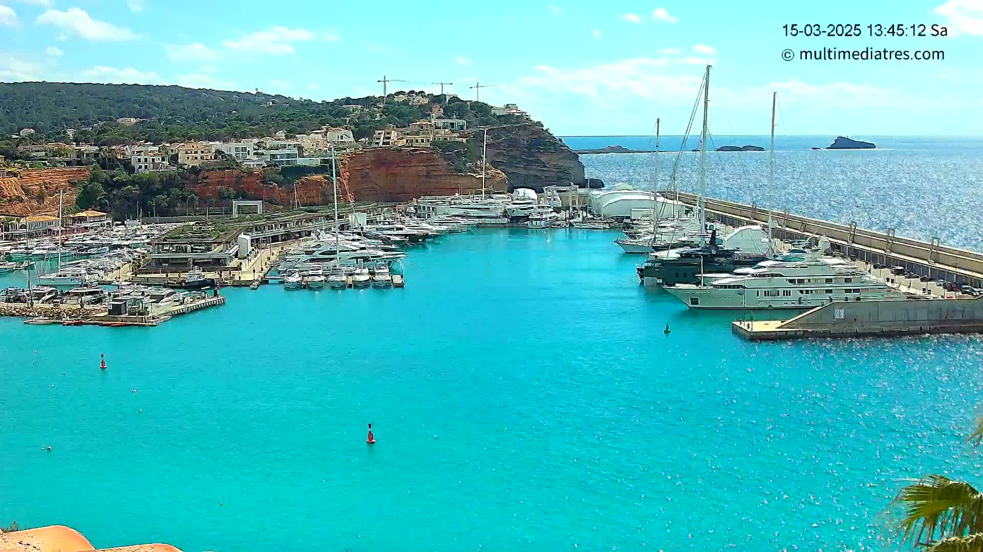 A marina filled with numerous yachts and boats is situated on bright turquoise water under a mostly sunny sky, with a red cliffside and buildings in the background.