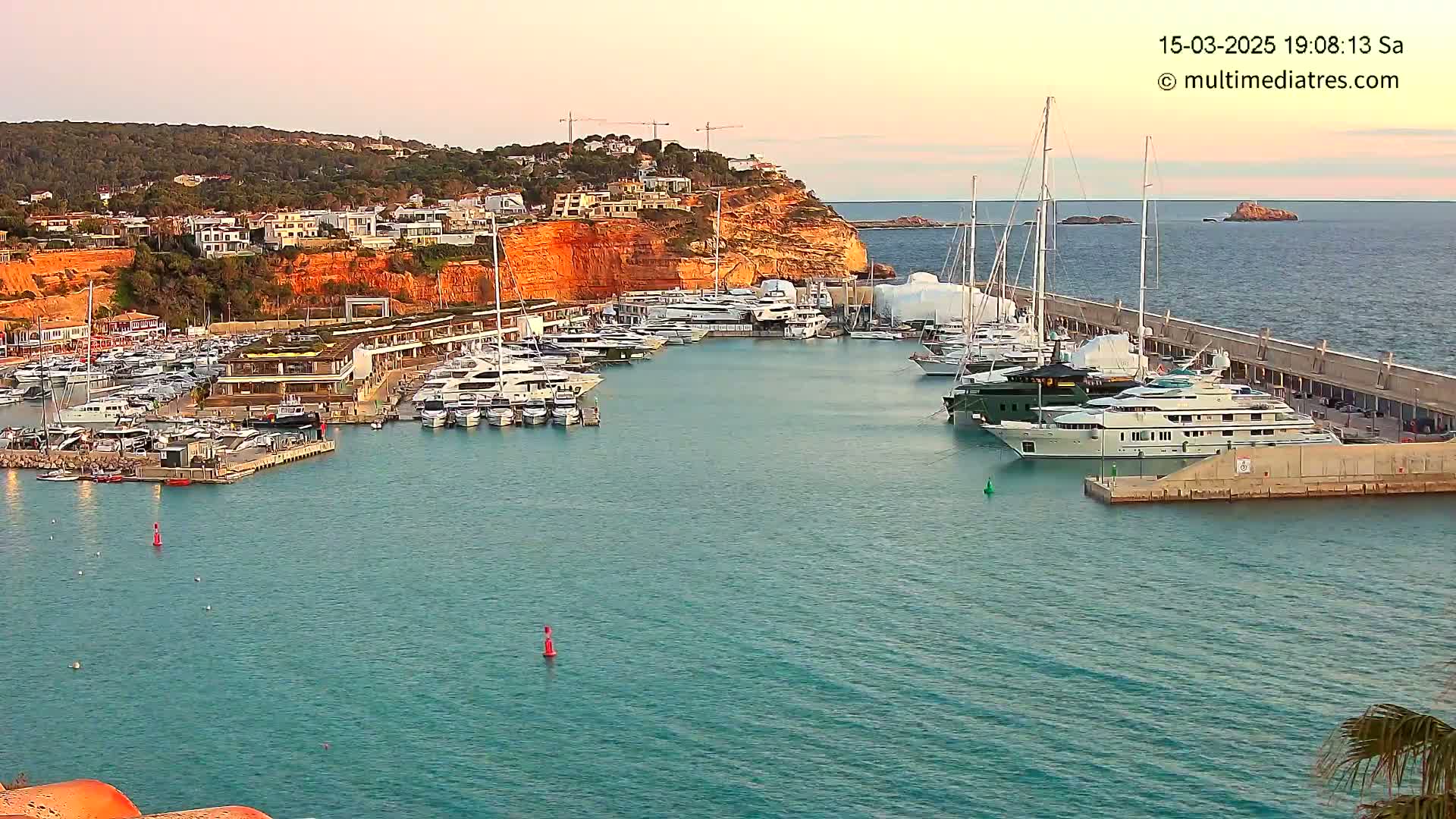 A marina filled with numerous yachts and boats is situated in a calm bay, at sunset, against a backdrop of a hillside town.