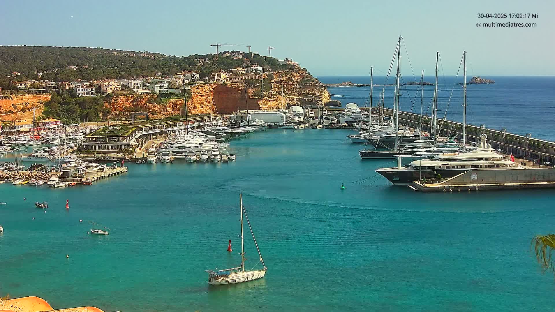 A marina filled with numerous yachts and sailboats is nestled in a calm, turquoise bay next to a red cliff lined with buildings under a clear, sunny sky.