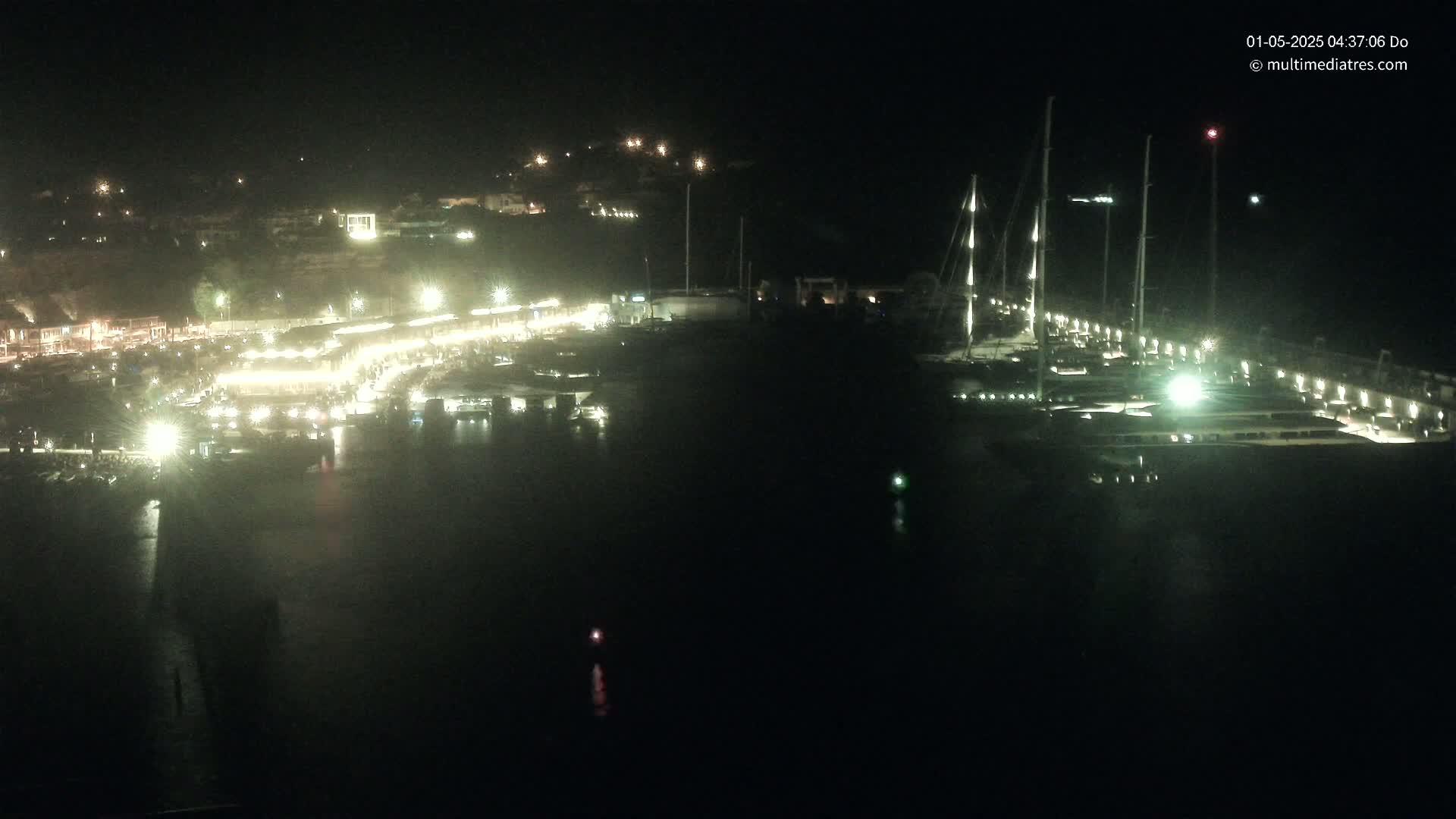 A nighttime, aerial view of a marina filled with boats, illuminated by lights along a waterfront with buildings on a hillside in the background.