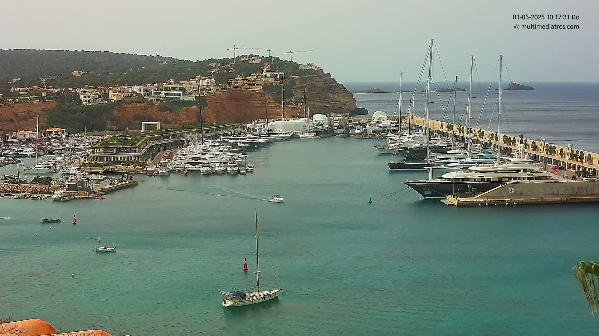 A marina filled with numerous yachts and sailboats is nestled in a calm, teal bay next to a hillside of buildings under an overcast sky.
