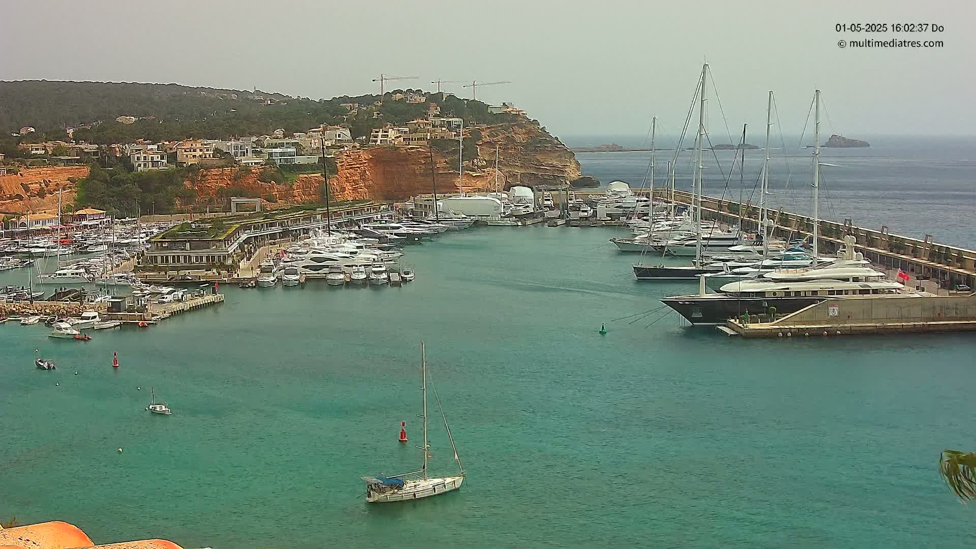 A marina filled with numerous yachts and sailboats is situated in a calm, teal-colored bay, under an overcast sky.