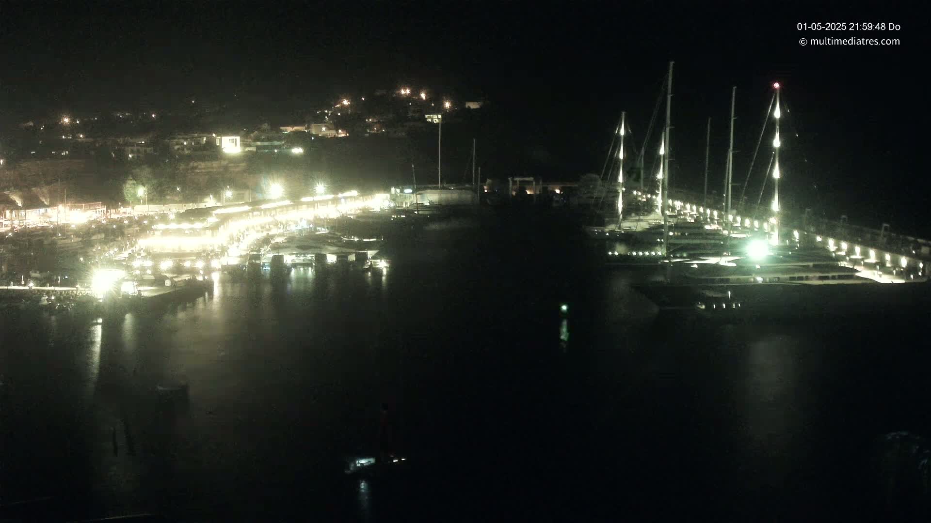 A nighttime view of a marina filled with boats, illuminated by lights along the docks and buildings on the hillside.