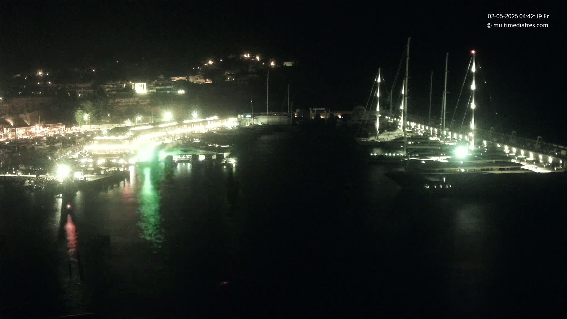 A nighttime view of a marina filled with docked sailboats, illuminated buildings along a shoreline, and a dark body of water reflecting the lights.