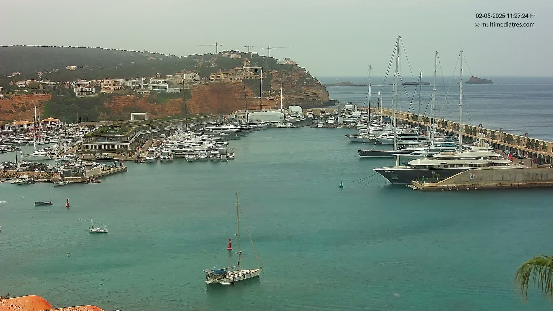 A marina filled with numerous yachts and sailboats is situated along a coastline, with overcast skies and calm water.