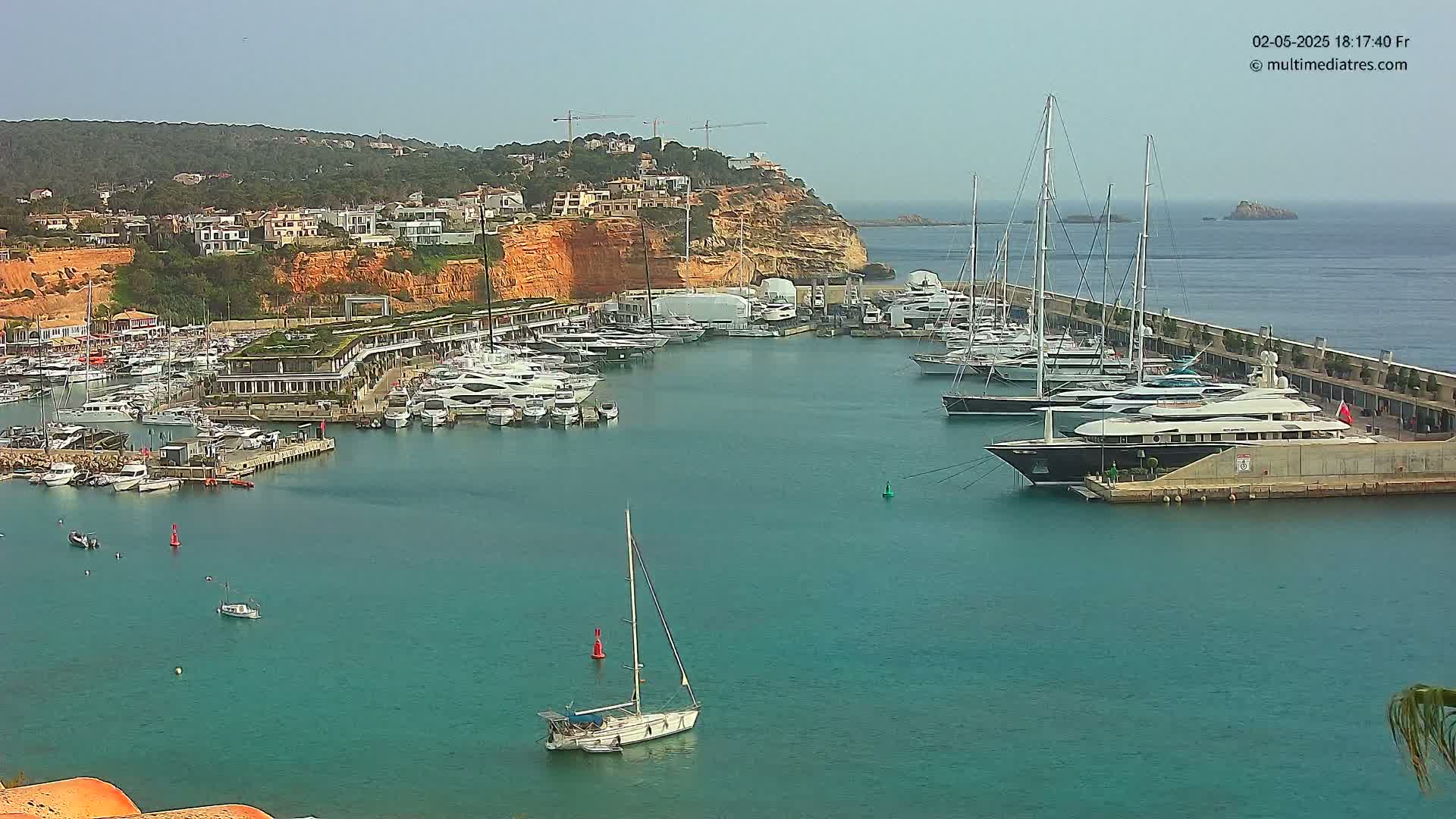 A marina filled with numerous yachts and sailboats is situated along a coastline on a sunny day.