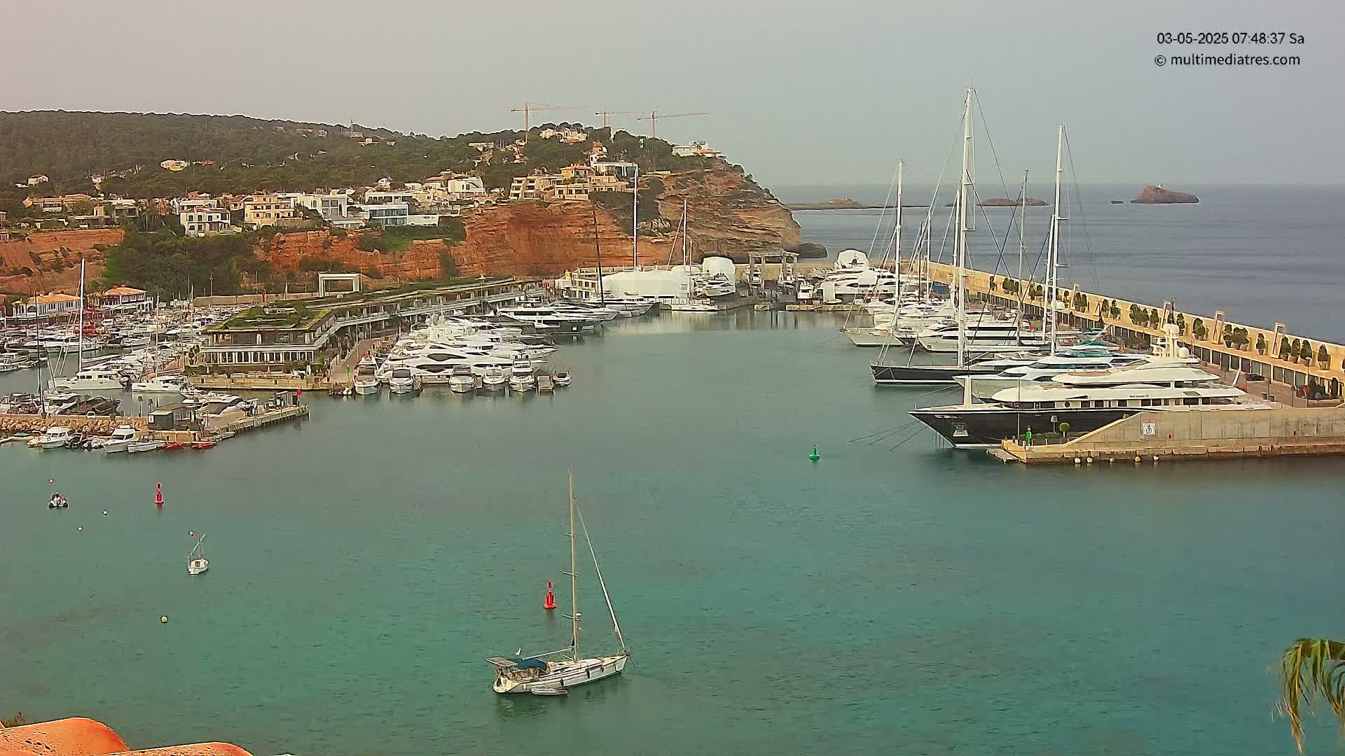 A marina filled with numerous yachts and sailboats is nestled against a reddish-brown cliffside under an overcast sky.