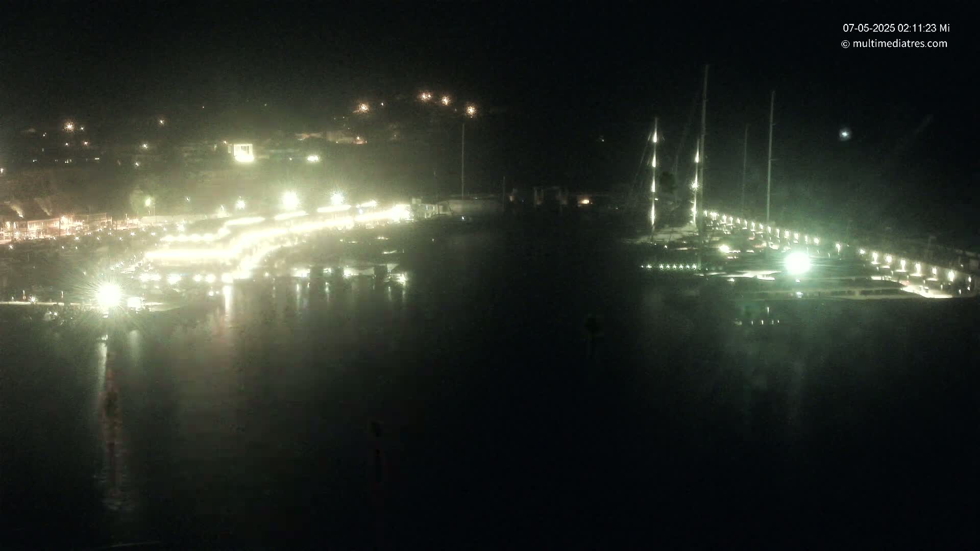 A nighttime harbor scene shows a marina with many boats and brightly lit docks, reflecting on calm water.