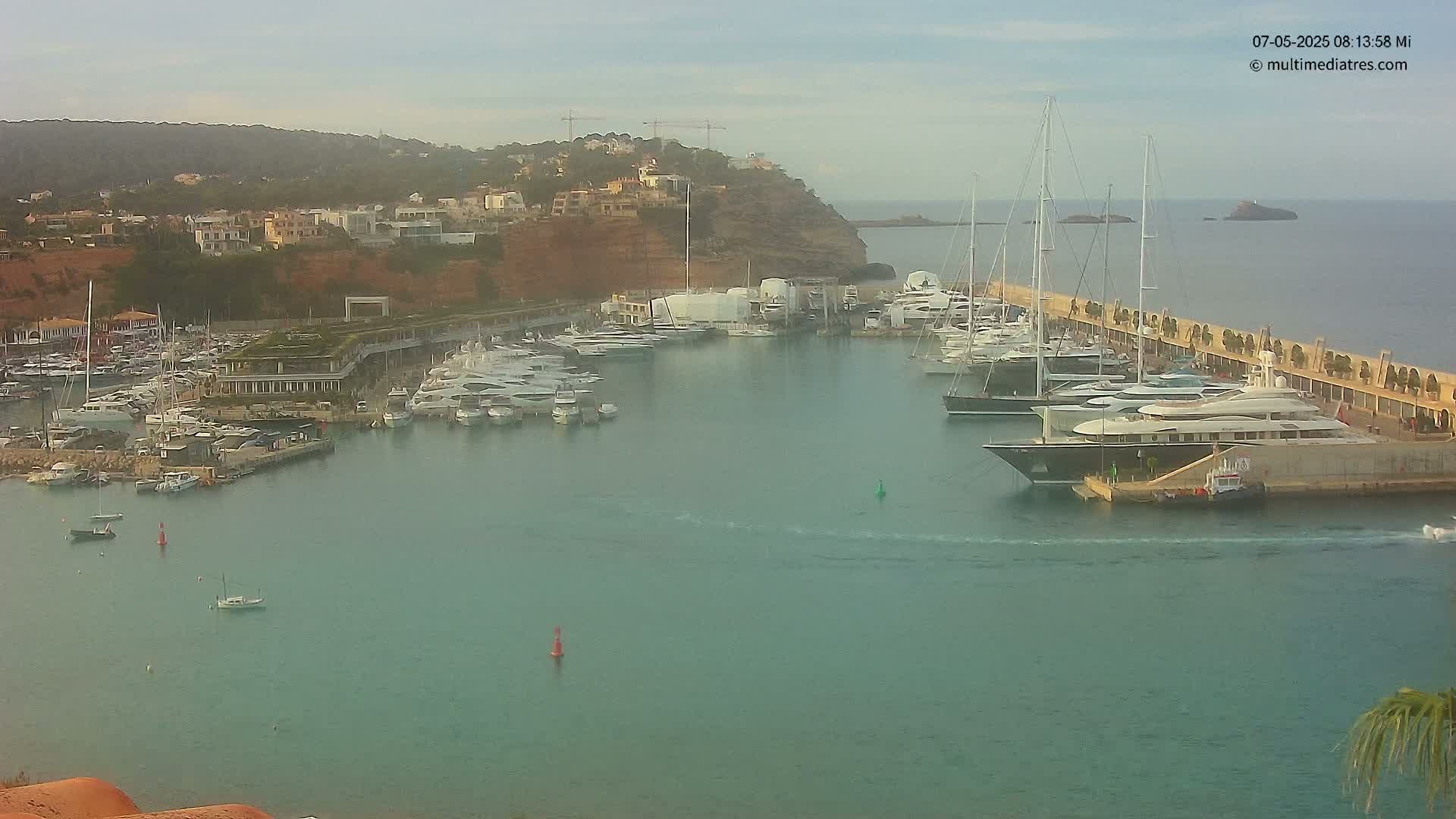 A marina filled with yachts and sailboats is nestled into a calm, teal-colored bay under a hazy sky.