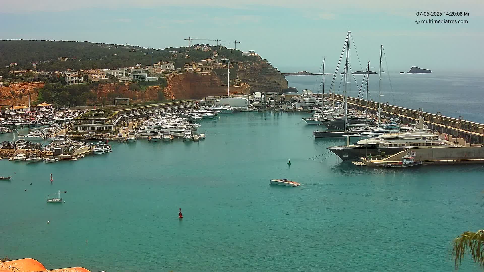 A marina filled with numerous yachts and boats is nestled into a calm, turquoise bay, backed by a hillside of buildings under a clear sky.