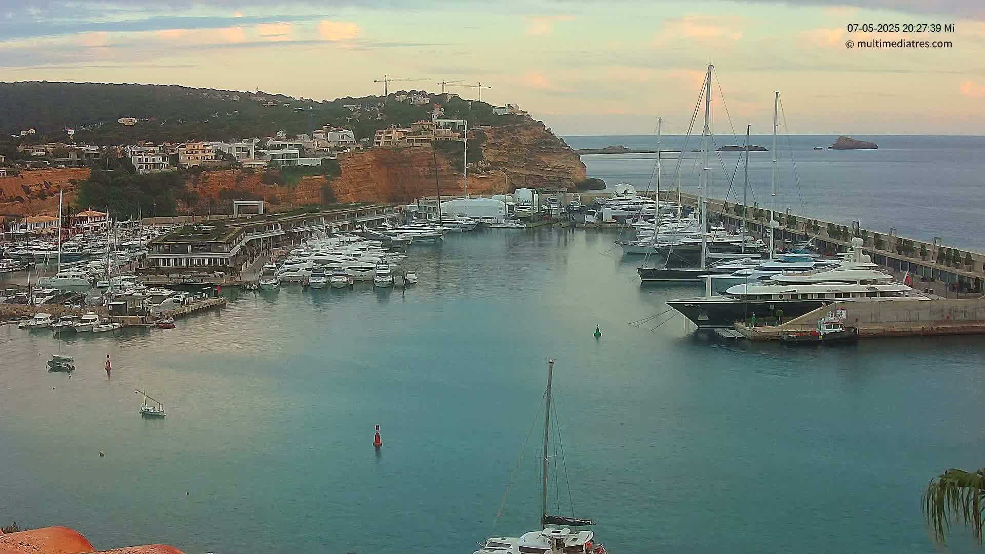A marina filled with many yachts and sailboats is nestled against a hillside of buildings under a partly cloudy sky.
