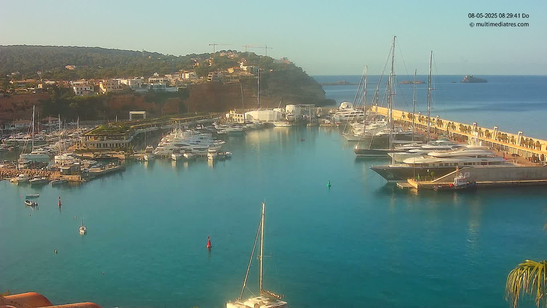 A marina filled with numerous yachts and sailboats is nestled against a hillside of buildings under a clear, sunny sky.