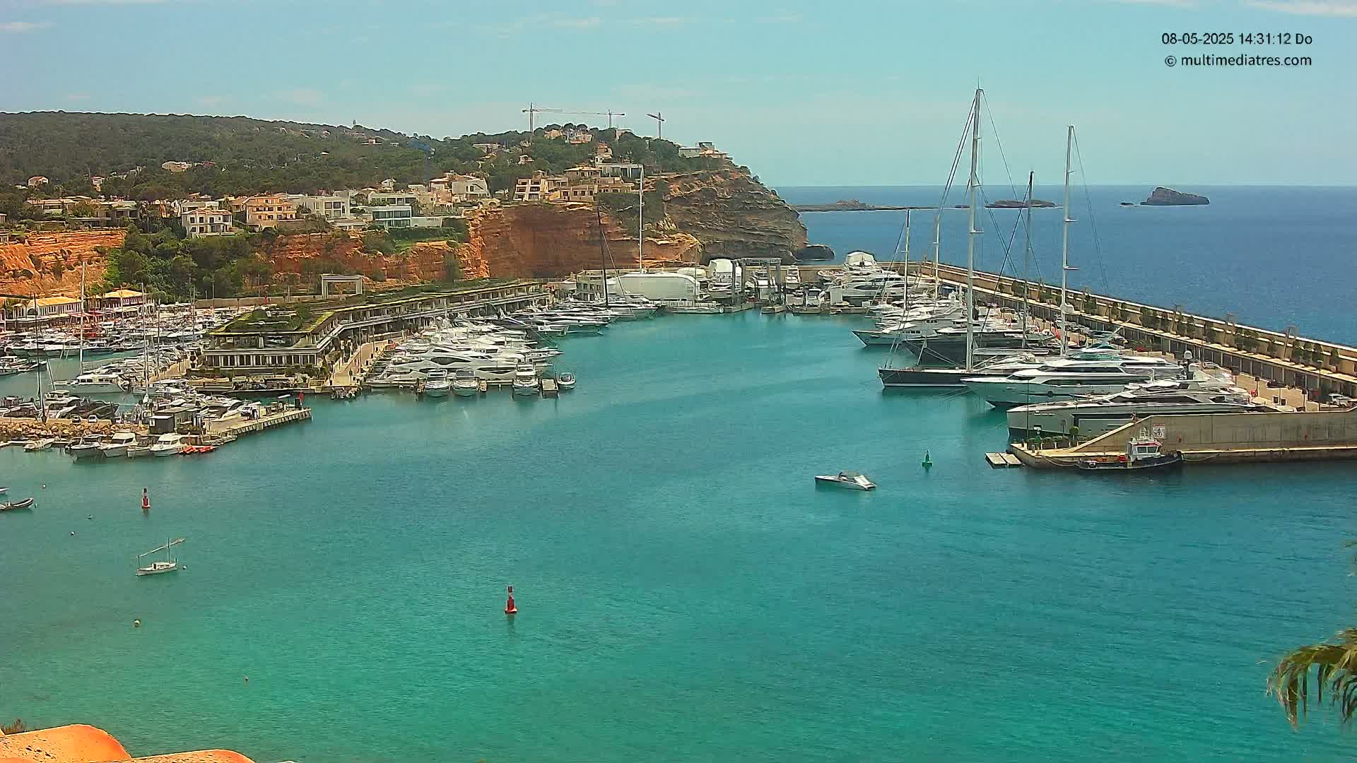 A marina filled with yachts and boats is nestled against a cliffside of buildings under a sunny sky.