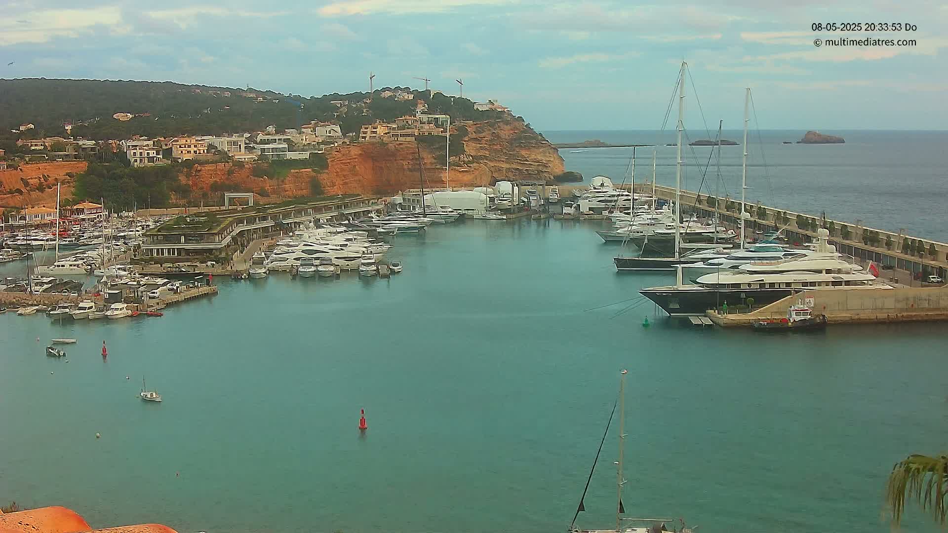 A marina filled with numerous yachts and boats is nestled against a cliffside town under a partly cloudy sky.