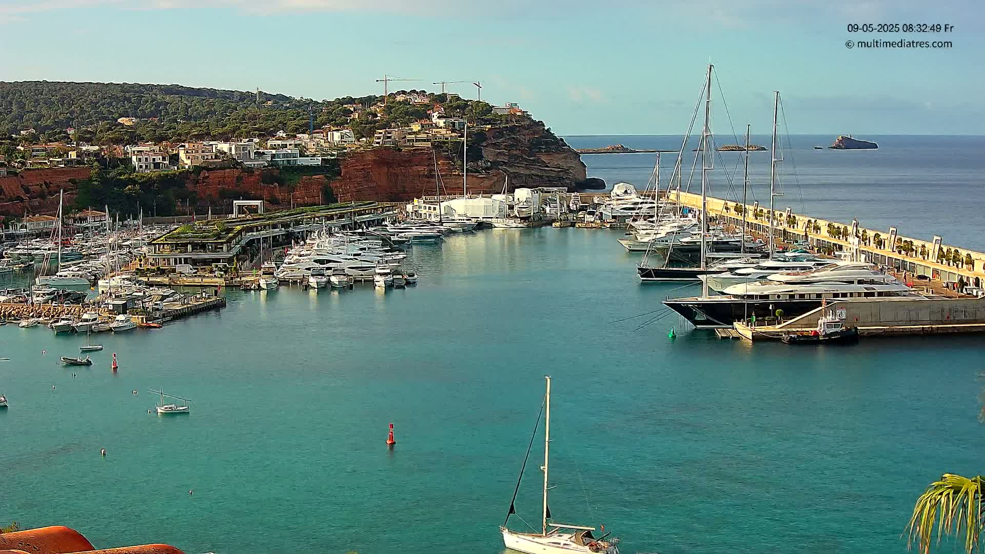A marina filled with numerous yachts and boats is situated along a rocky coastline under a clear sky.