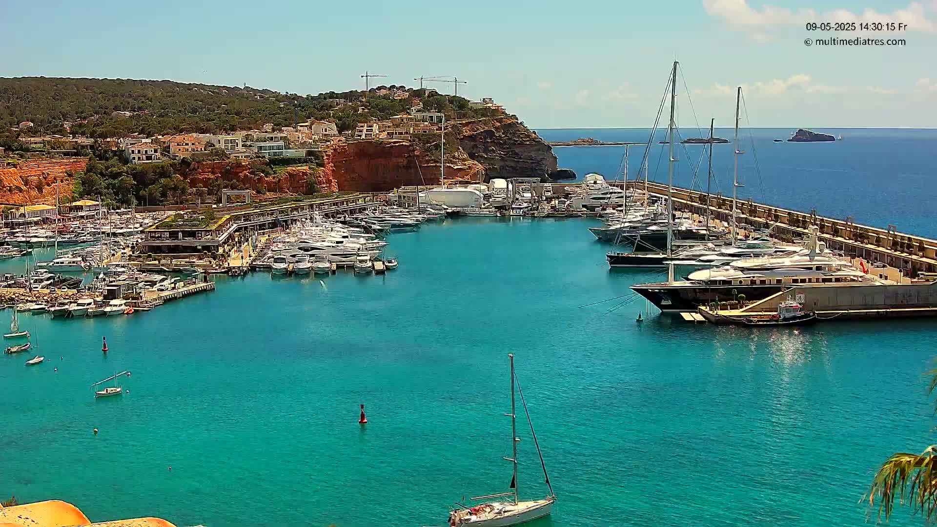 A marina filled with numerous yachts and boats is nestled into a turquoise bay on a sunny day, with a red-cliffed hillside rising behind it.