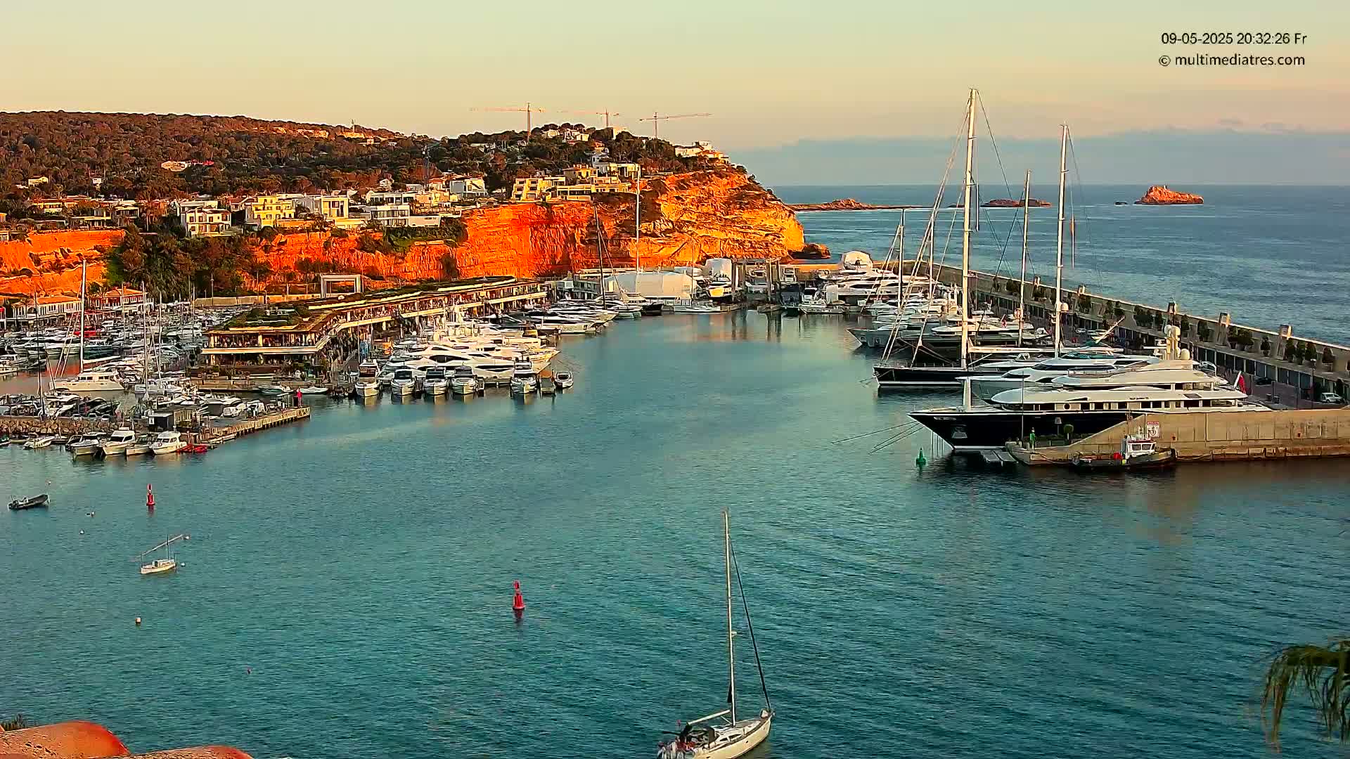 A marina filled with numerous yachts and sailboats is situated along a cliffside, under a clear evening sky.
