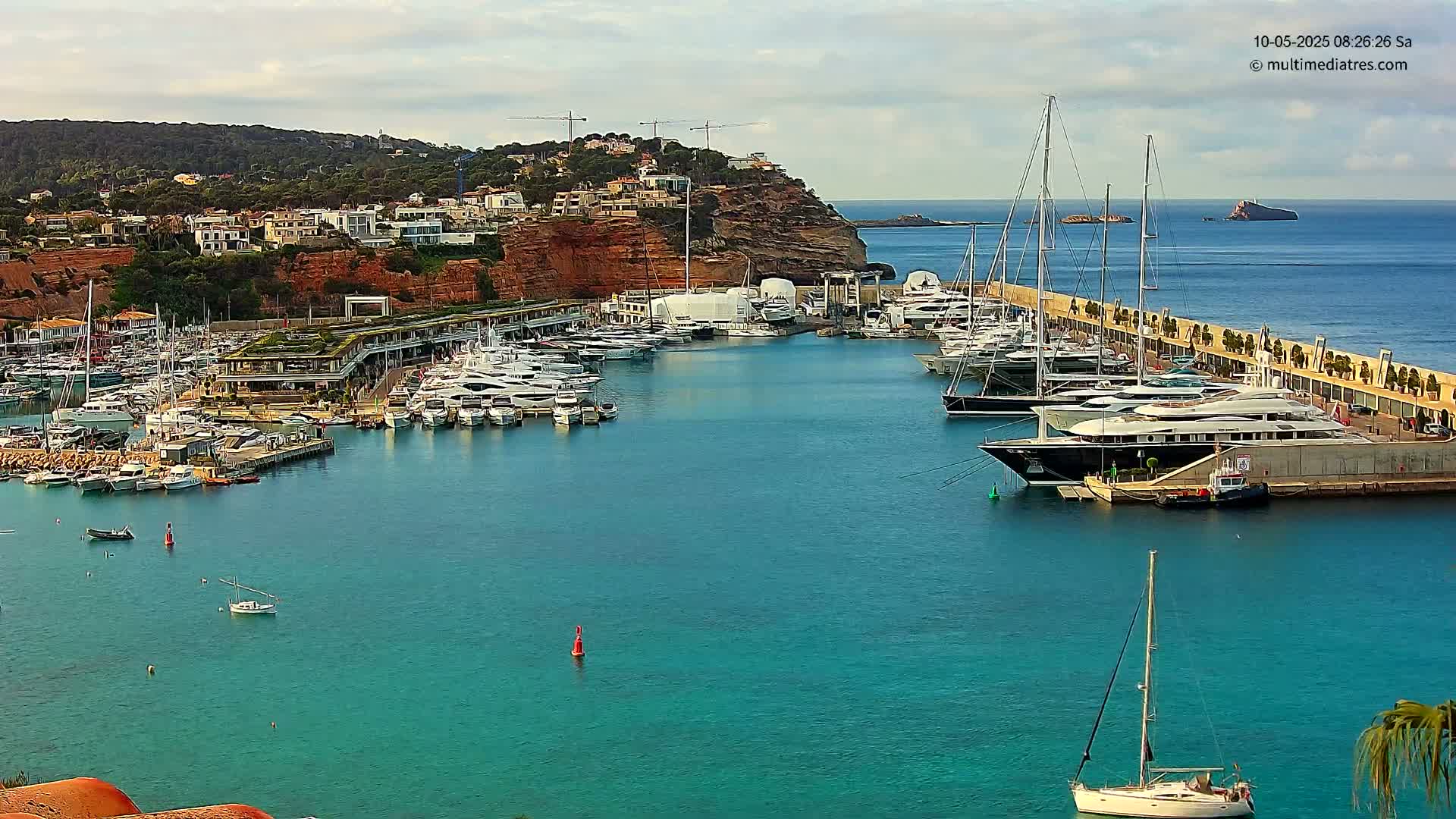A marina filled with numerous yachts and boats is nestled against a red cliffside, under a partly cloudy sky.