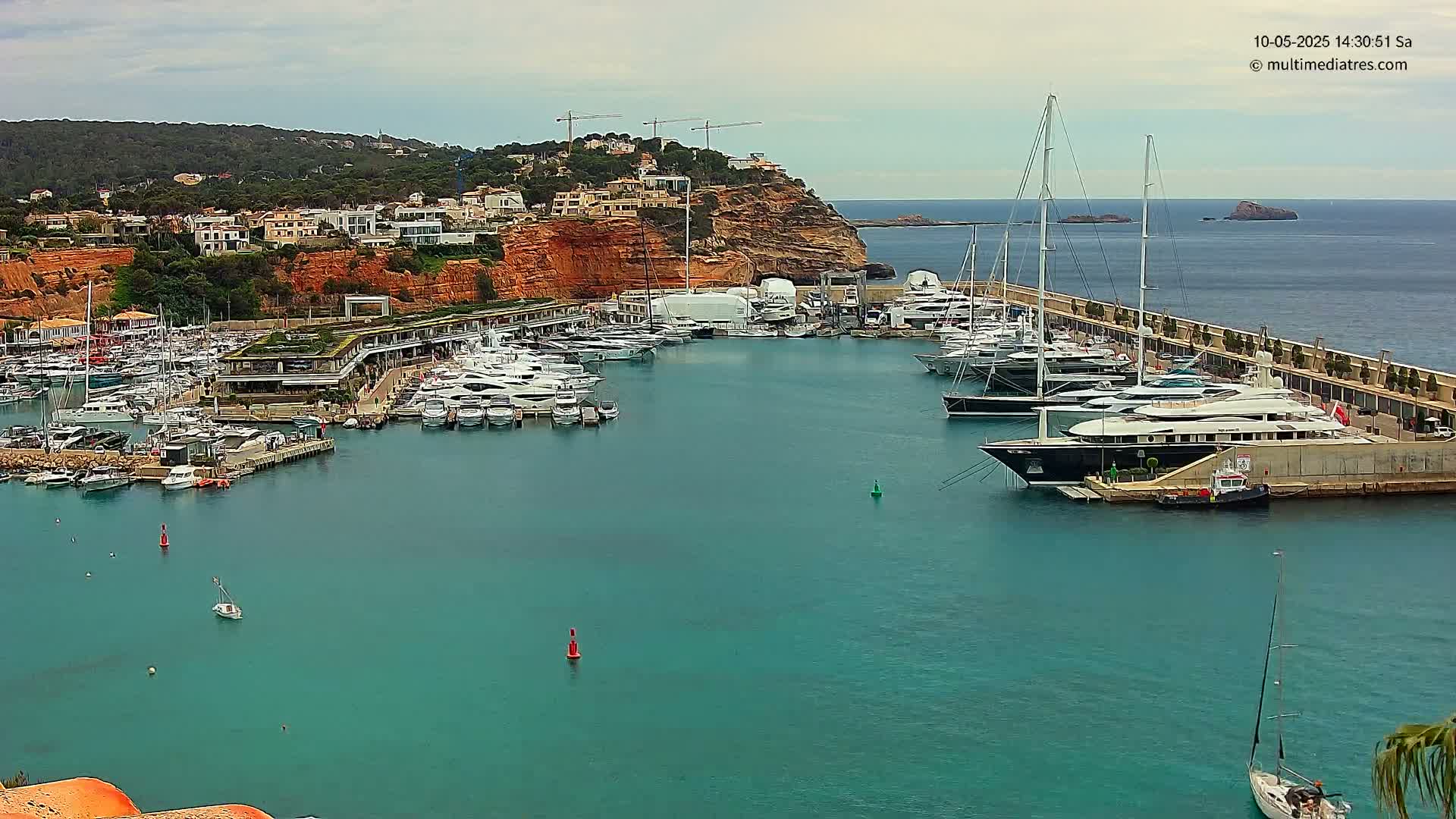 A marina filled with numerous yachts and boats is nestled against a red cliffside under a mostly cloudy sky.