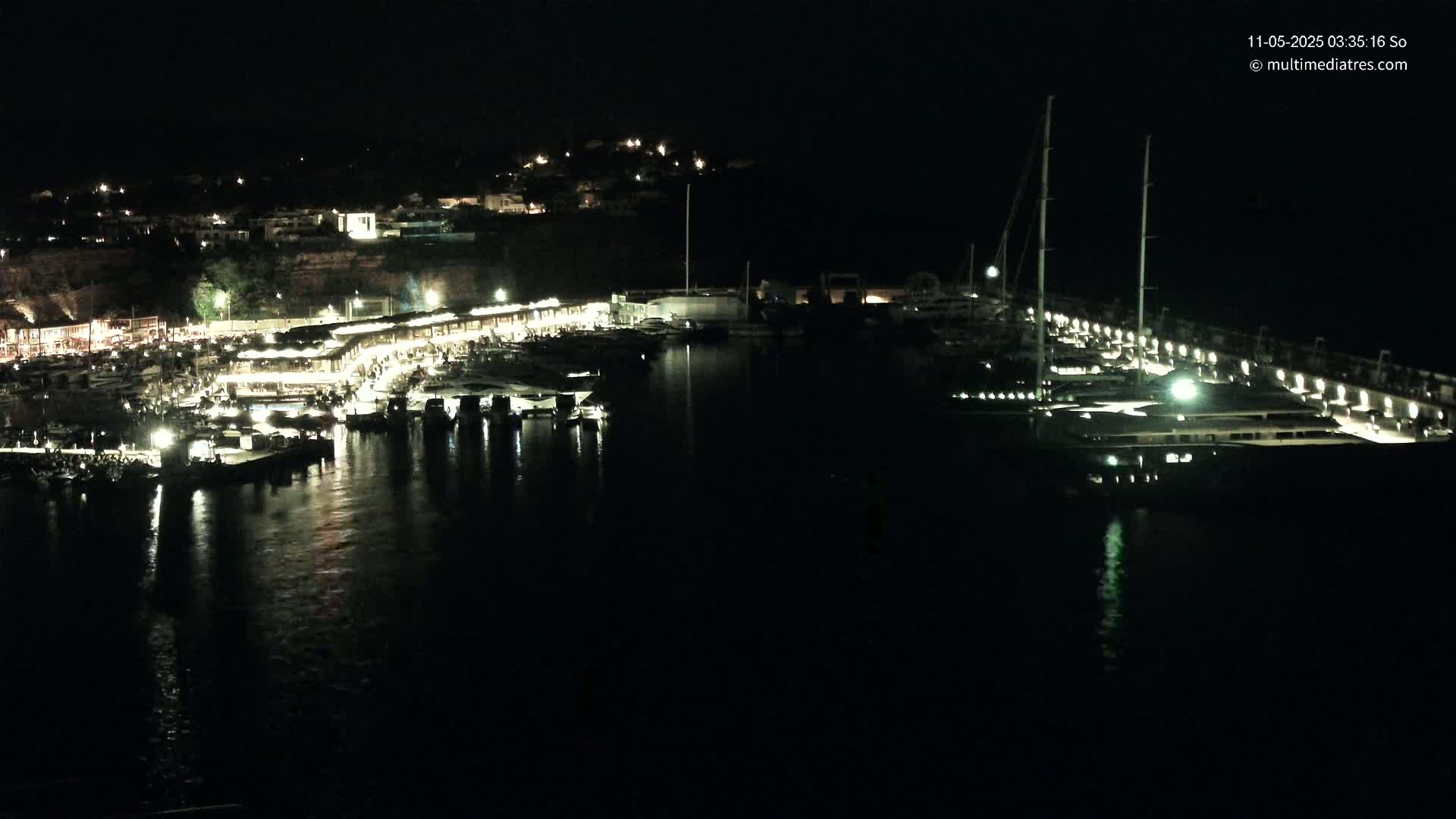 A nighttime view of a harbor filled with boats, lit buildings along the shore, and a hill in the background.