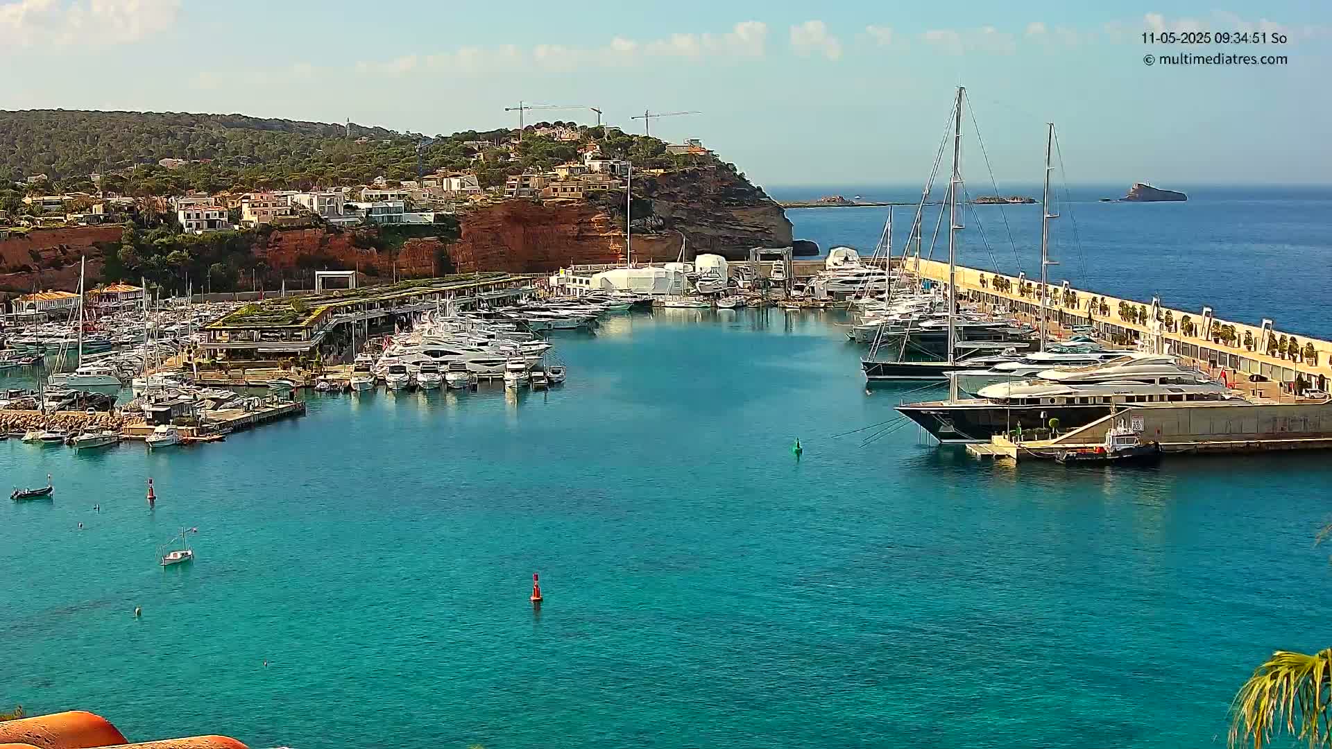 A marina filled with numerous yachts and boats is nestled in a turquoise bay under a clear, sunny sky, with a hillside of buildings in the background.