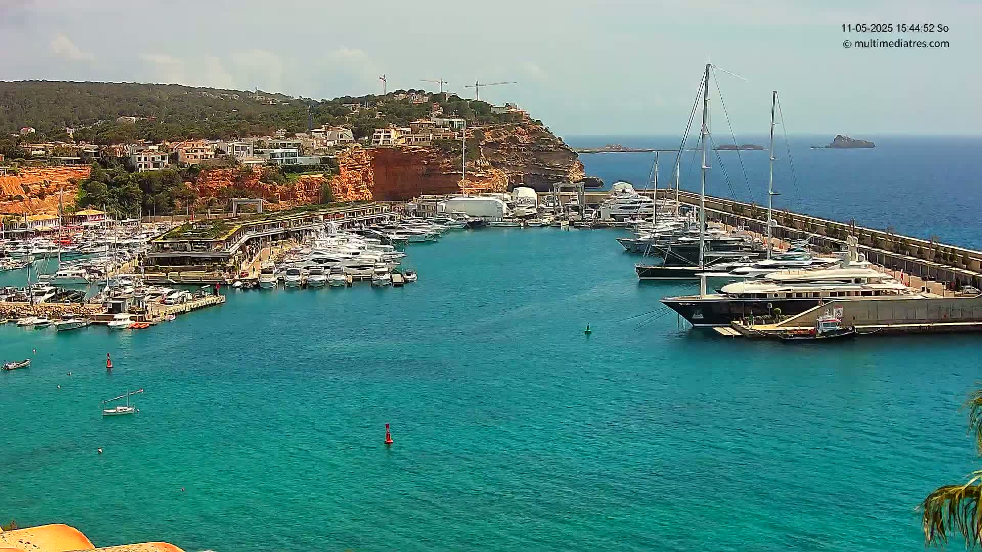 A marina filled with numerous yachts and boats is situated in a calm, turquoise bay against a backdrop of red-cliffed hills under a clear sky.