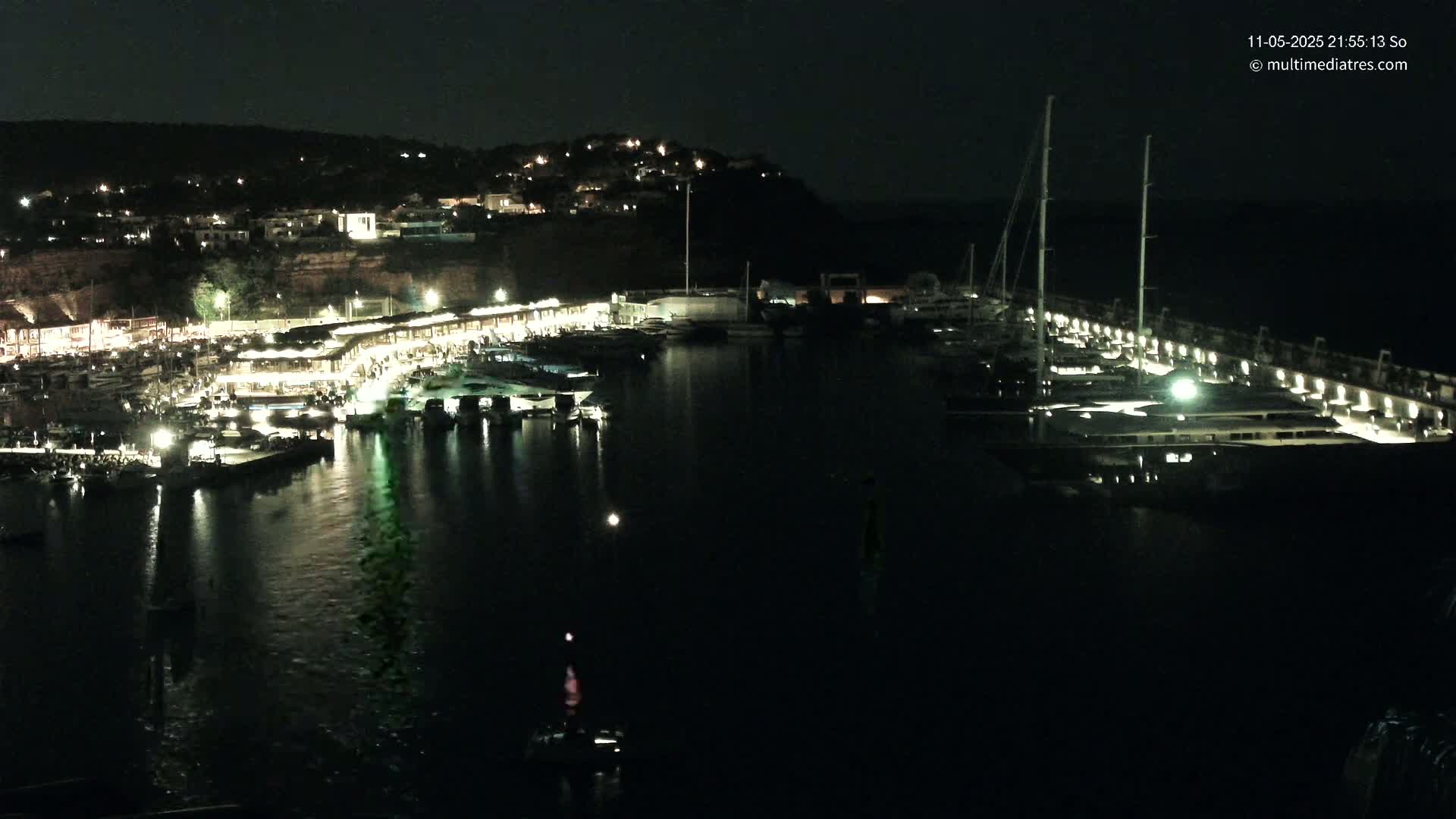 A nighttime view of a harbor filled with boats, docked alongside a lit-up pier, with a hillside of buildings visible in the background.