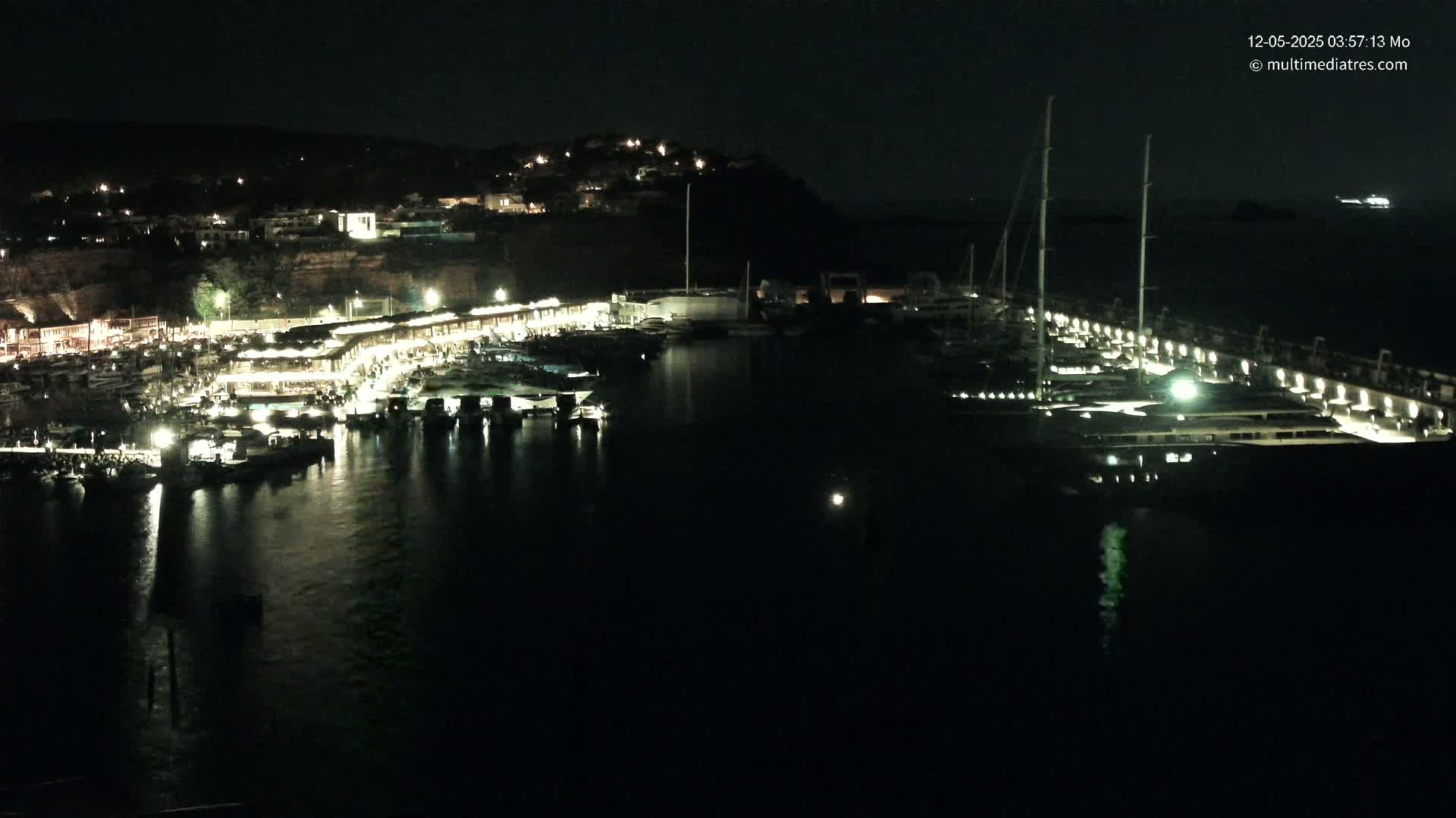 A nighttime aerial view shows a marina filled with boats and lit buildings along a dark, calm body of water, with a hill of houses visible in the background.