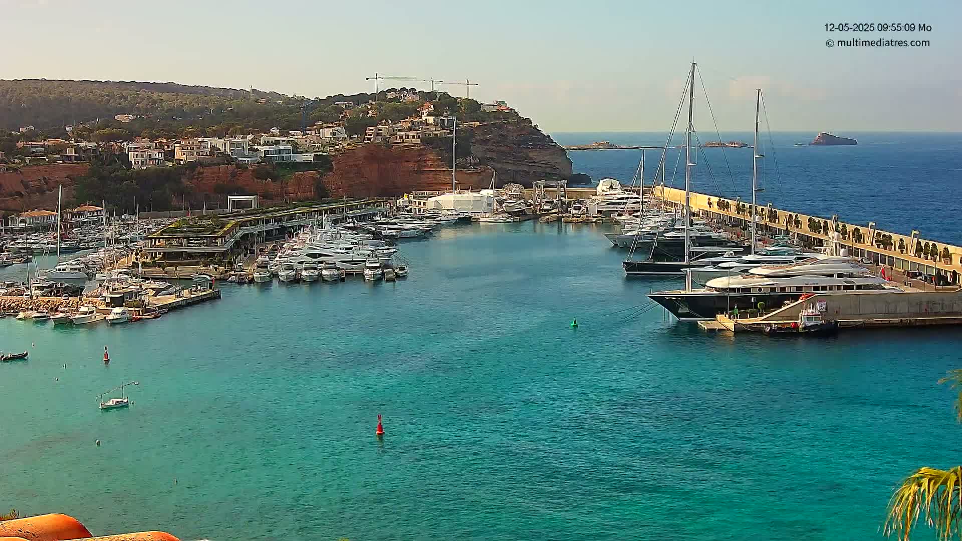 A marina filled with numerous yachts and boats is situated in a calm, clear turquoise body of water, with a residential area and reddish cliffs visible in the background under a bright, sunny sky.