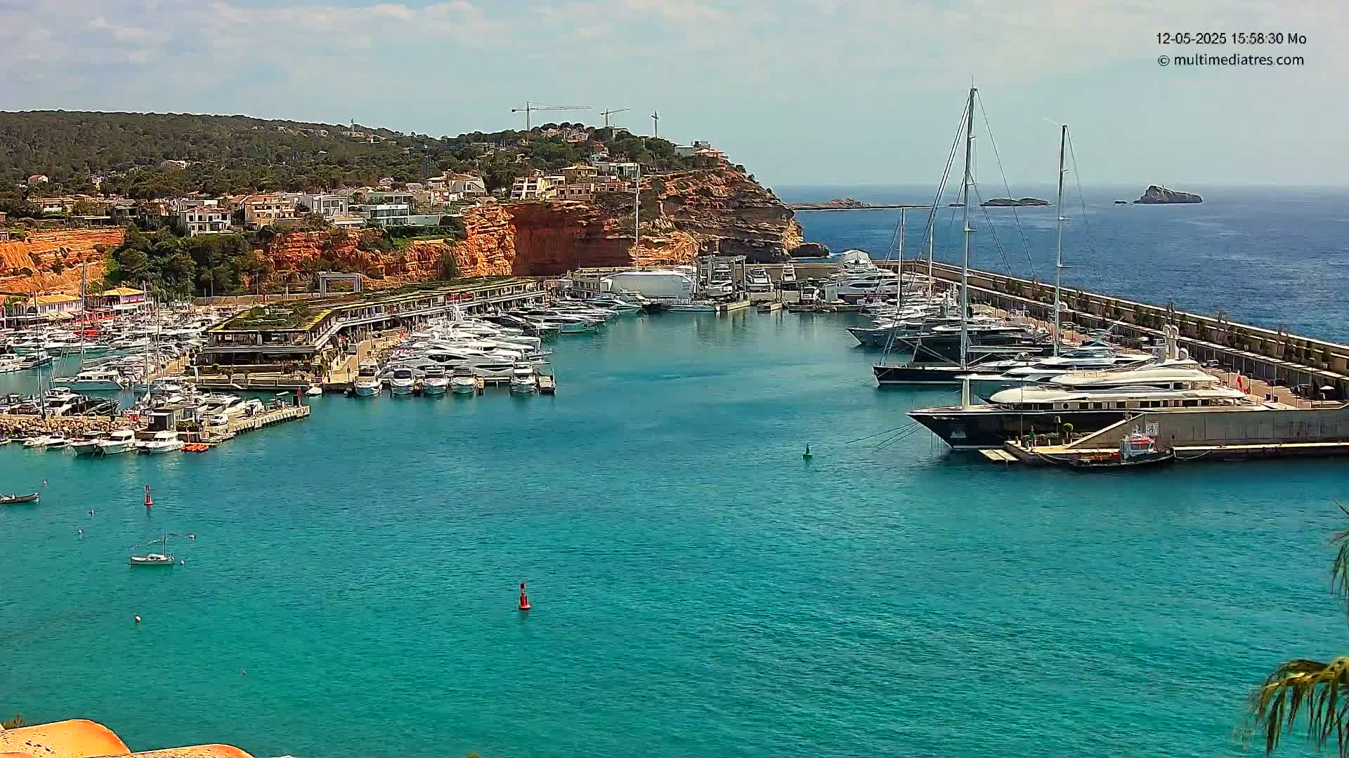 A marina filled with numerous yachts and boats is situated on a calm, turquoise body of water next to a red cliffside with buildings and vegetation.