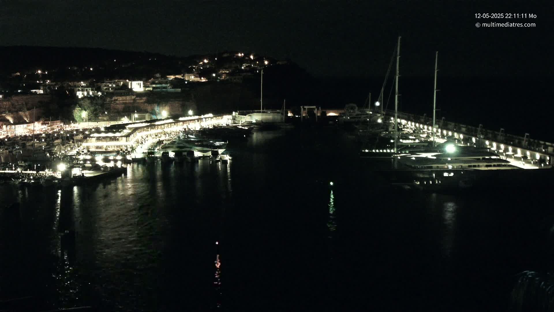 A nighttime view of a harbor filled with boats, illuminated buildings along the shore, and a hill in the background.