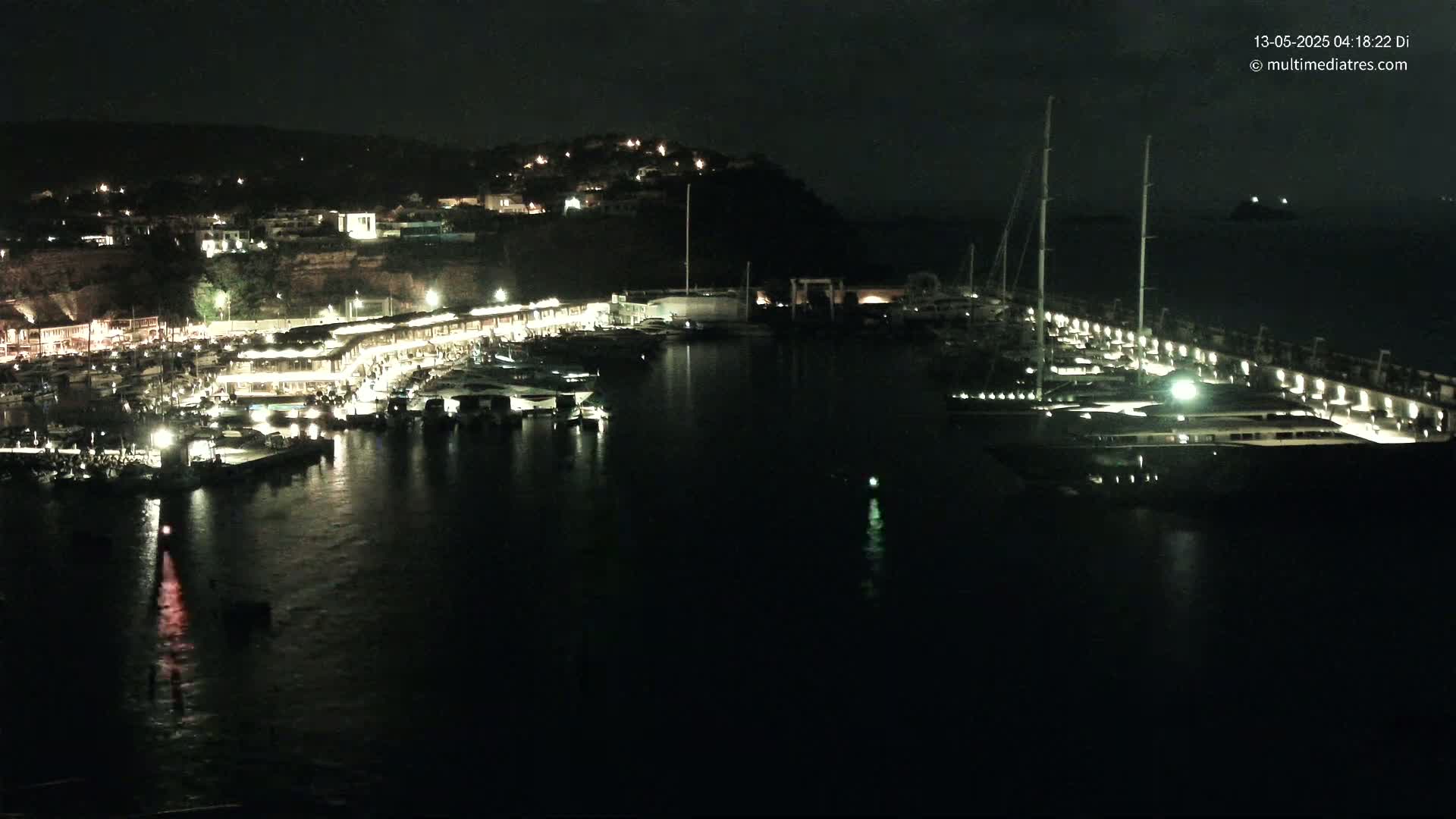A nighttime view of a harbor filled with boats, illuminated buildings along the shoreline, and a hill in the background.