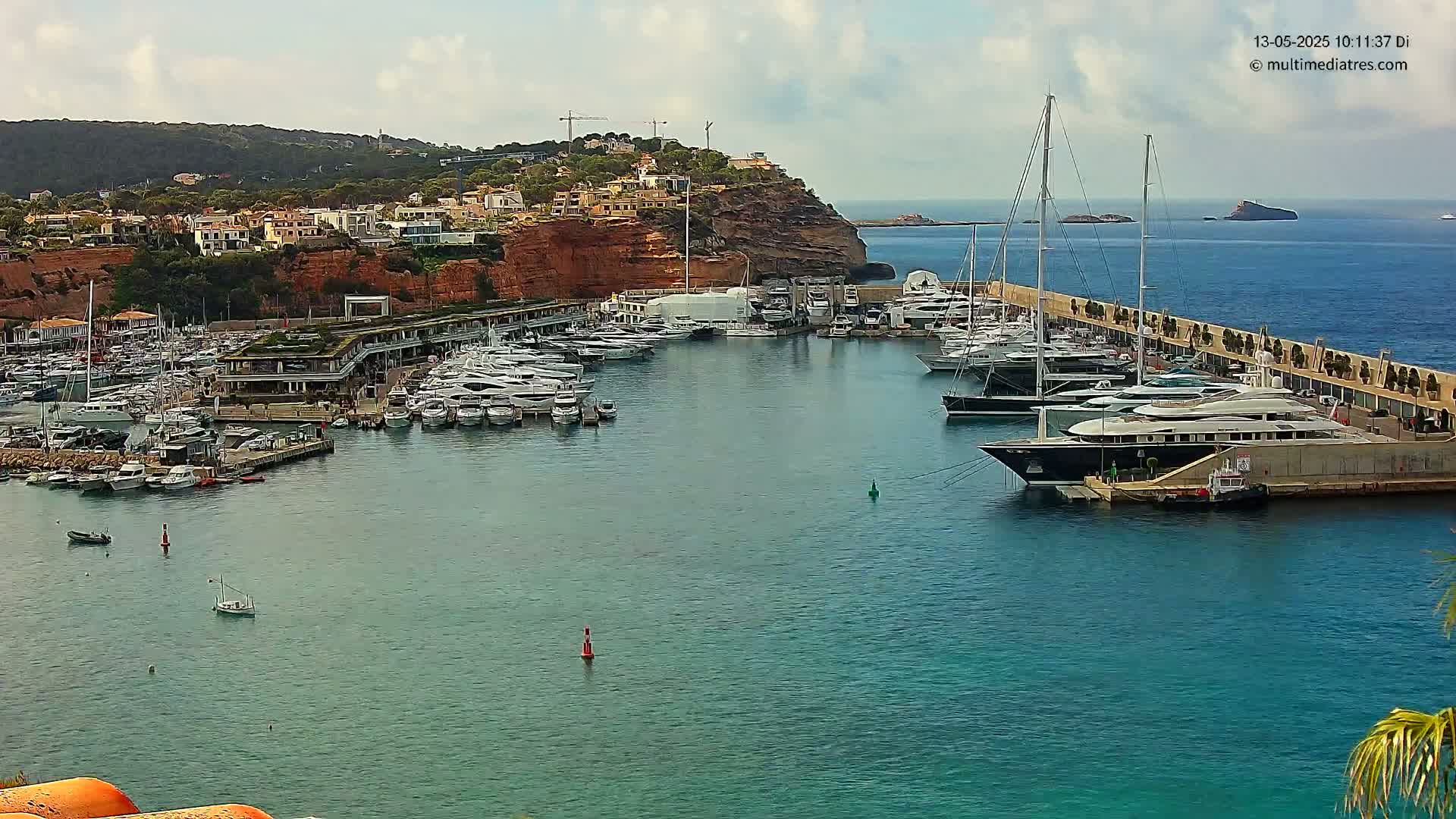 A marina filled with numerous yachts and boats is nestled against a rocky hillside under a partly cloudy sky.