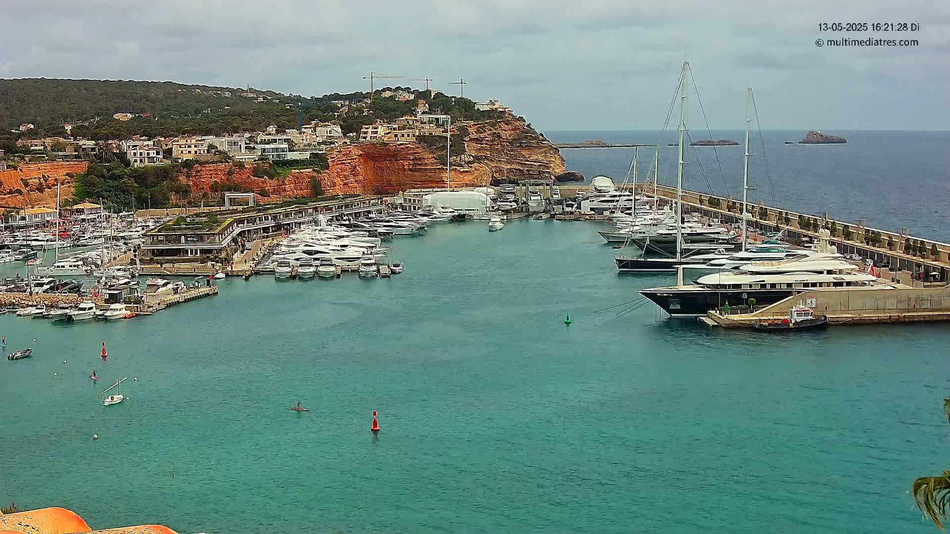 A marina filled with numerous yachts and boats is situated on a calm, teal body of water next to a red cliff, under an overcast sky.